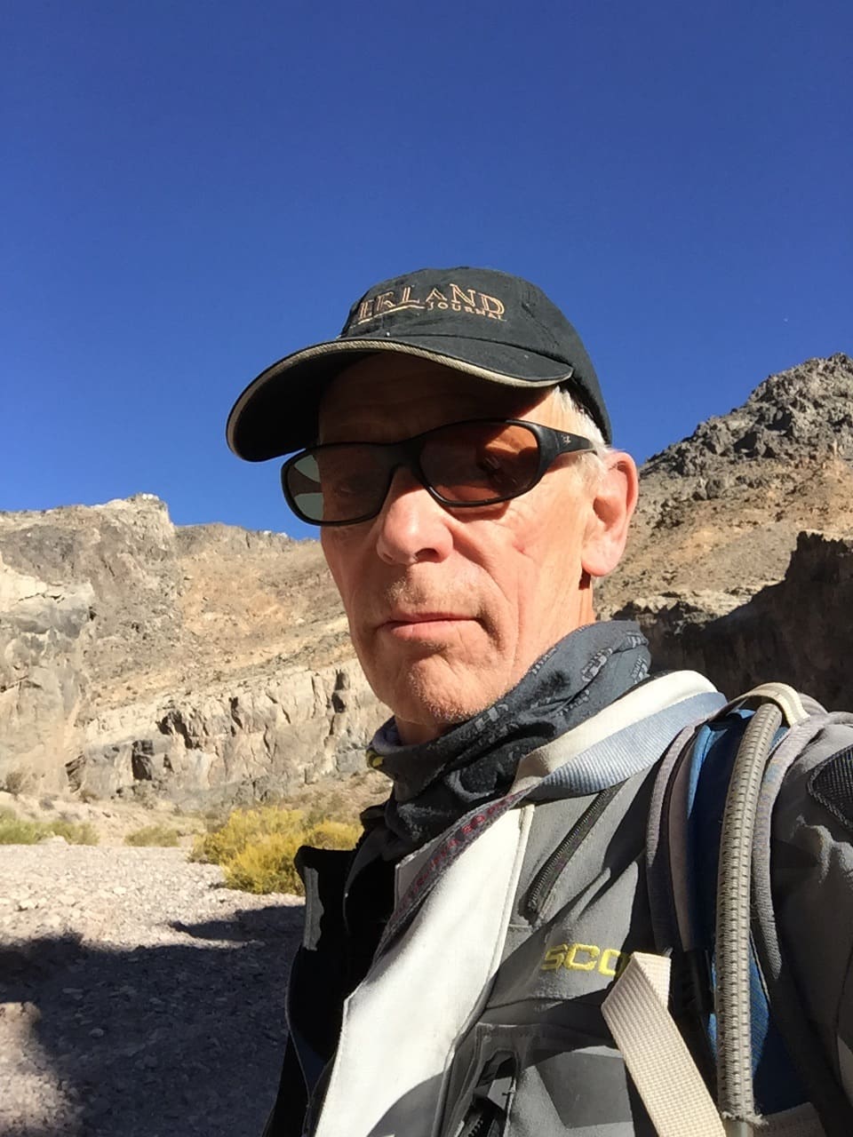 An older man wearing sunglasses, a hat, and outdoor gear stands in a rocky desert landscape under a clear blue sky, with cliffs and hills in the background—capturing the spirit of adventure embodied by Arden Kysely at the 2026 Celebration of Life.