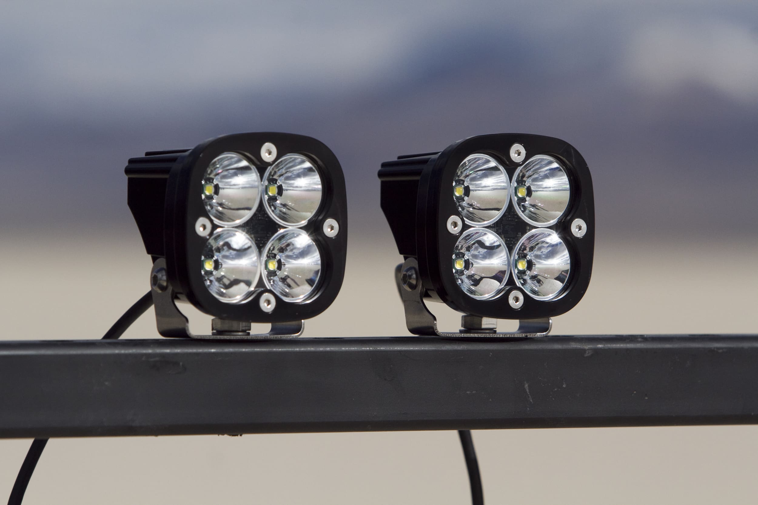 Two black, square-shaped LED driving lights, each with four circular bulbs, are mounted on a metal bar outdoors against a blurred background.