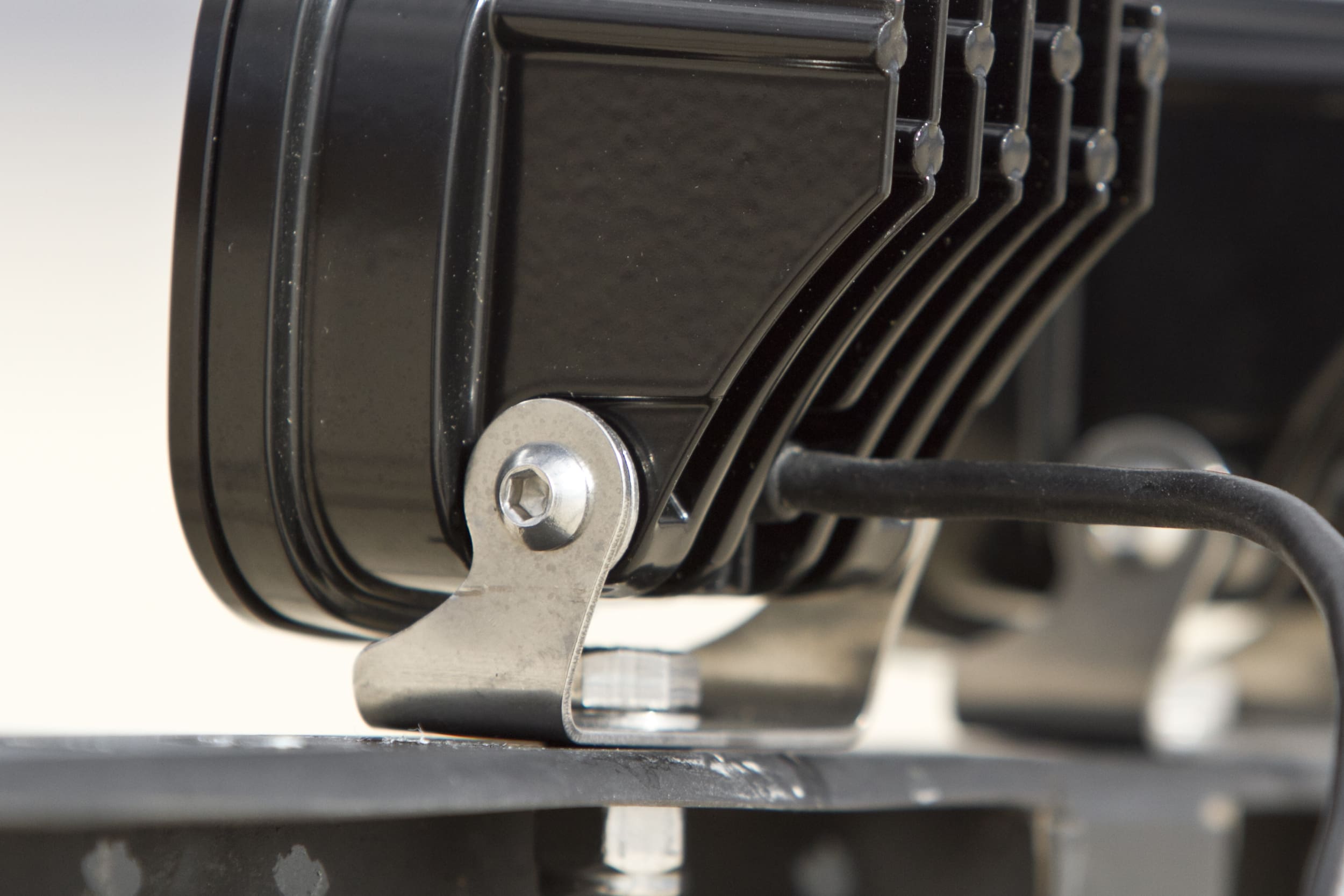 Close-up of a black metal bracket secured by a bolt, attached to a ribbed device with a cable connected to it, possibly part of driving lights or other electronic equipment.