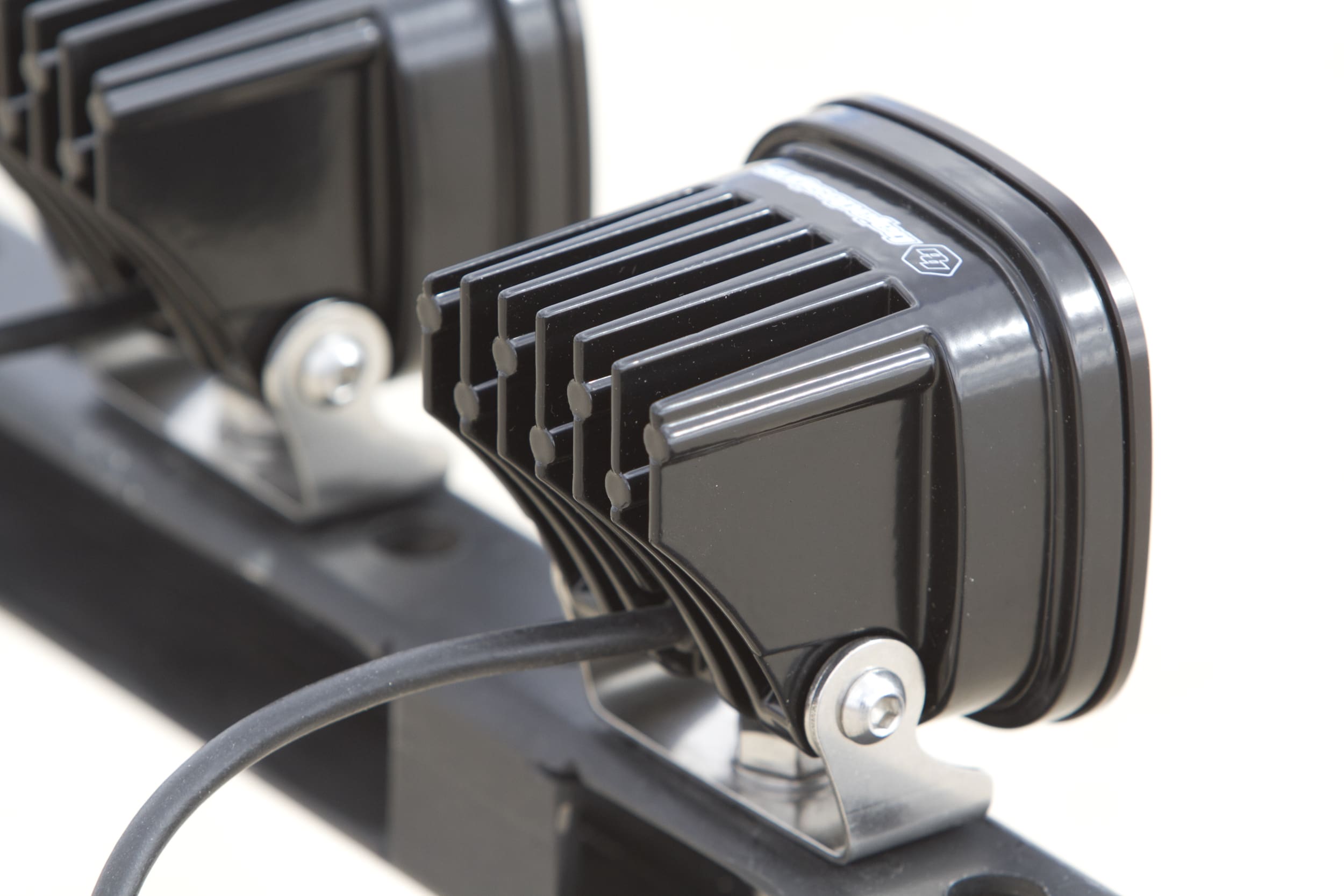 Close-up of two black rectangular LED driving lights with heat sink fins, mounted on a metal bracket. A black cable extends from each light. The background is out of focus.