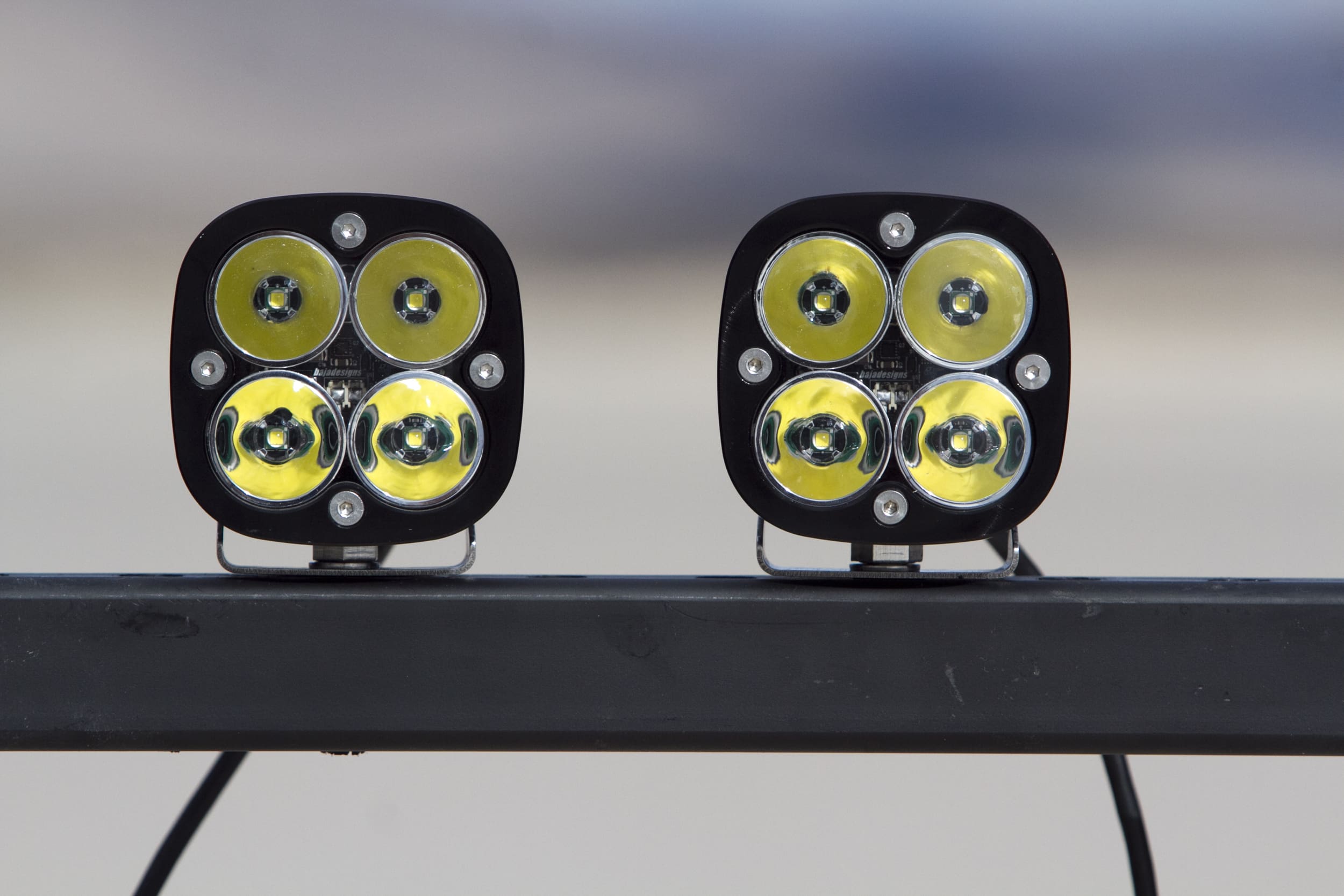 Two square-shaped LED off-road driving lights with four yellow bulbs each, mounted side by side on a metal bar, against a blurred background.
