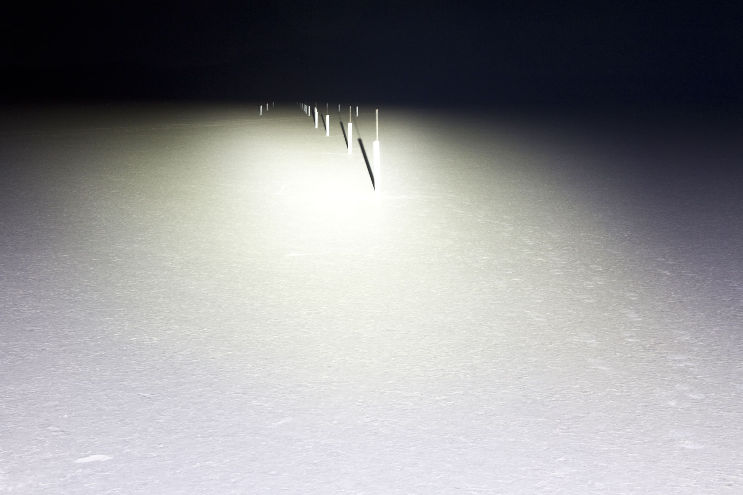 A row of thin poles stretches into the distance across a flat, snow-covered surface, illuminated by bright driving lights, with darkness in the background.