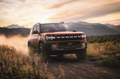 A red Ford Bronco with Integrated Camping Features drives on a dirt road through tall grass, kicking up dust with mountains and a dramatic sunset sky in the background.