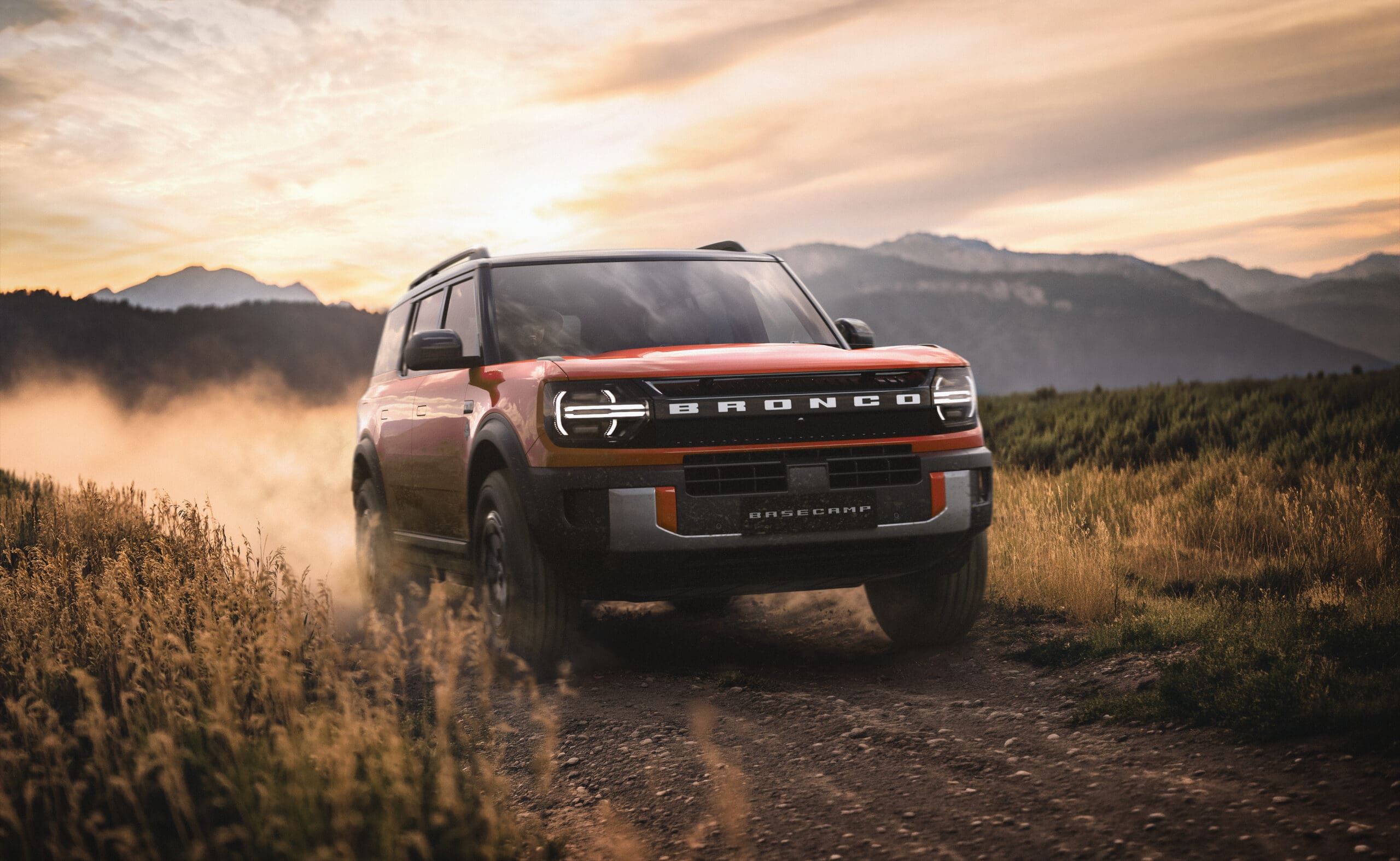A red Ford Bronco with Integrated Camping Features drives on a dirt road through tall grass, kicking up dust with mountains and a dramatic sunset sky in the background.