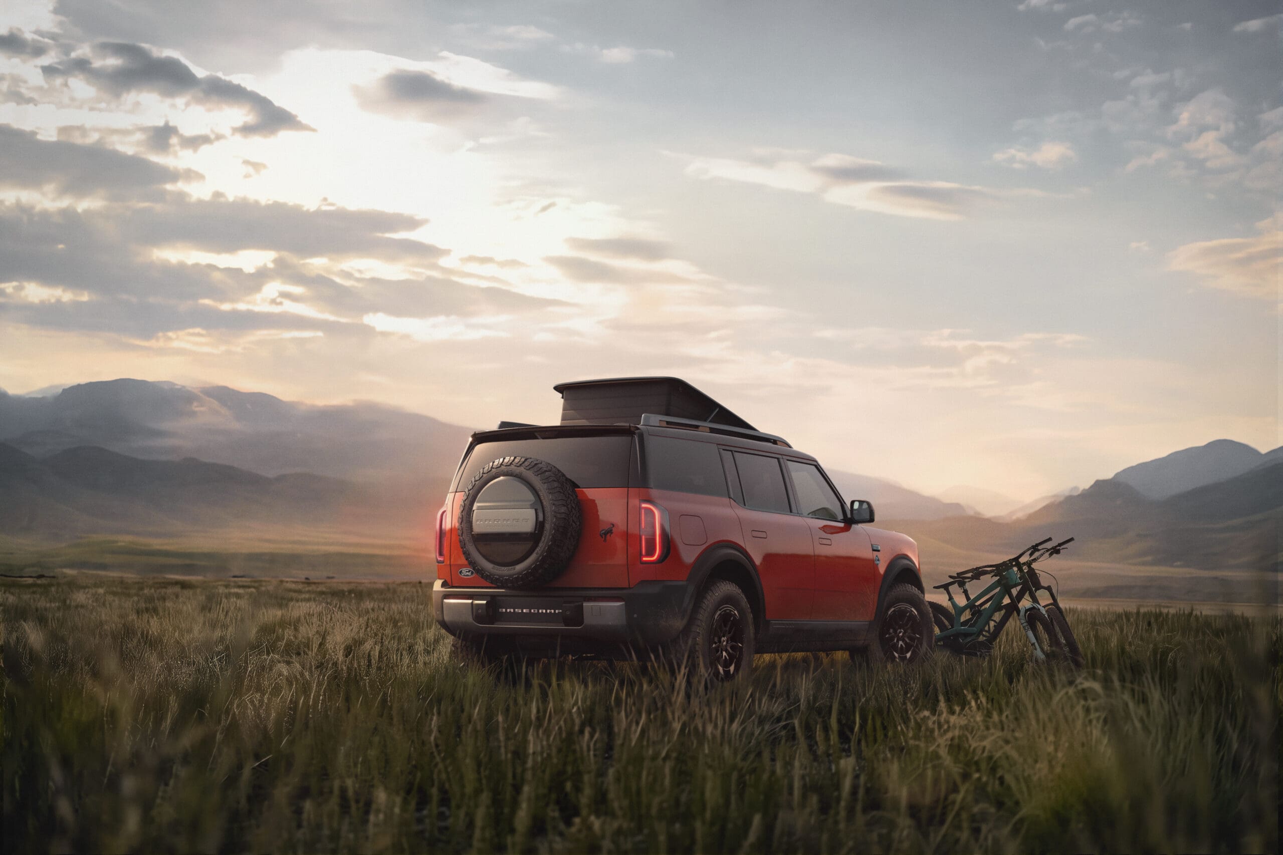 A red Ford Bronco SUV with a roof tent is parked in a grassy field near mountains at sunset. Two bicycles are leaning against the vehicle.