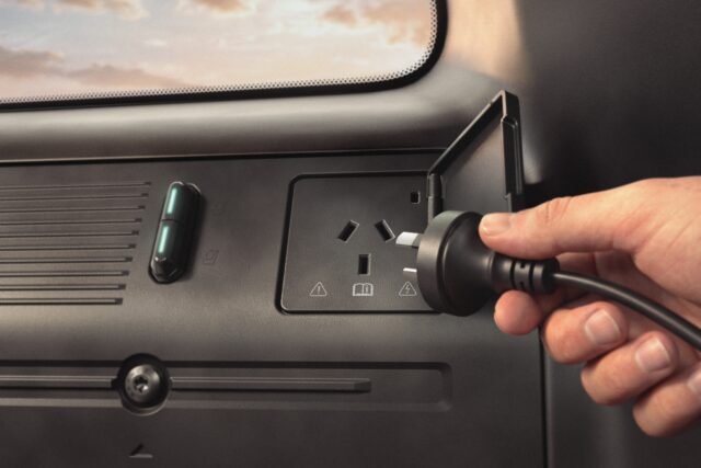 A hand is holding a plug near an electrical outlet inside a Ford Bronco, preparing to insert it. The outlet has warning symbols and a flipping cover.