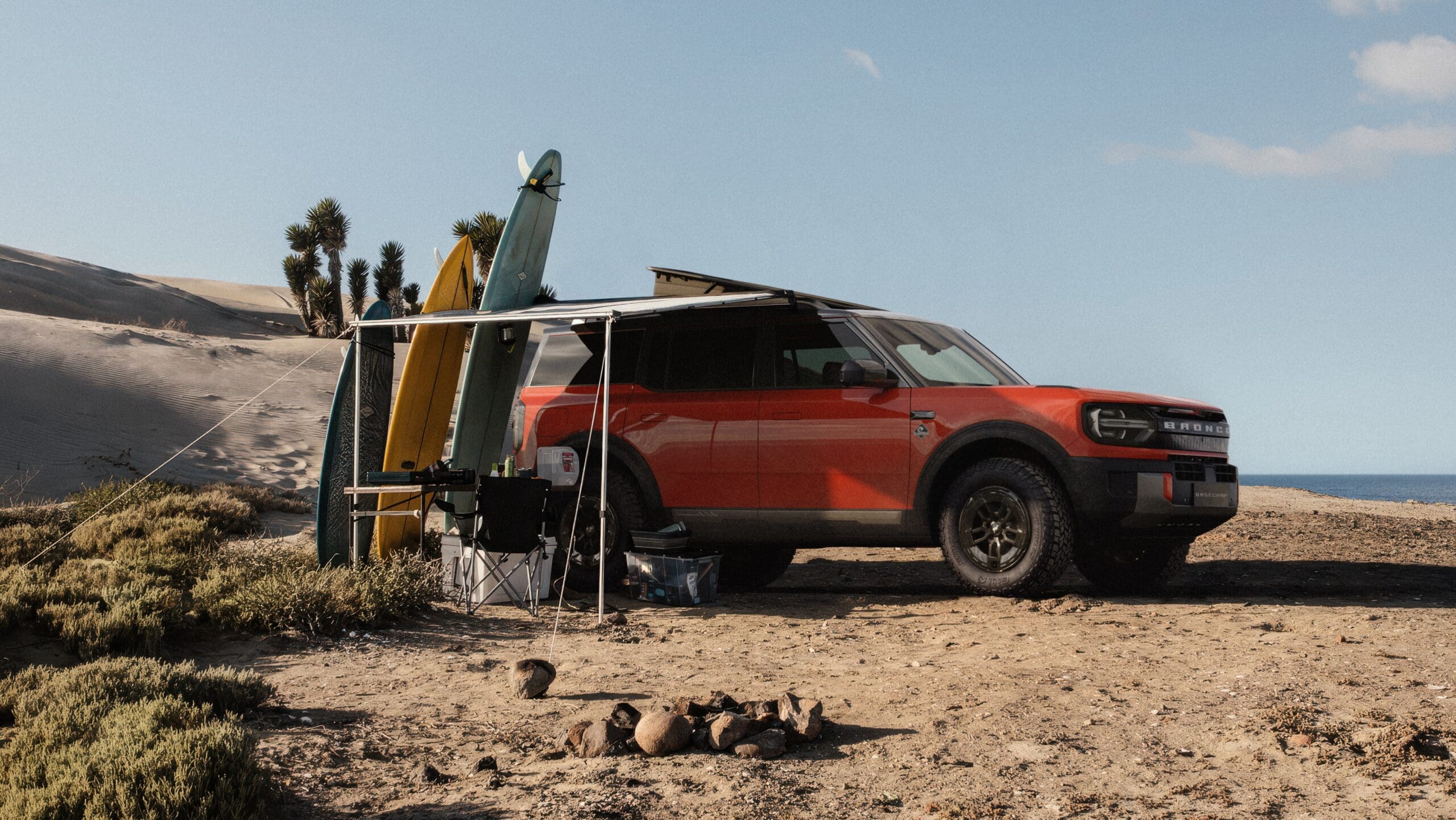 A red Ford Bronco SUV is parked on a sandy beach with several surfboards propped beside it. Camping gear is set up nearby, and a fire pit sits in the foreground by the dunes, sparse plants, and ocean.