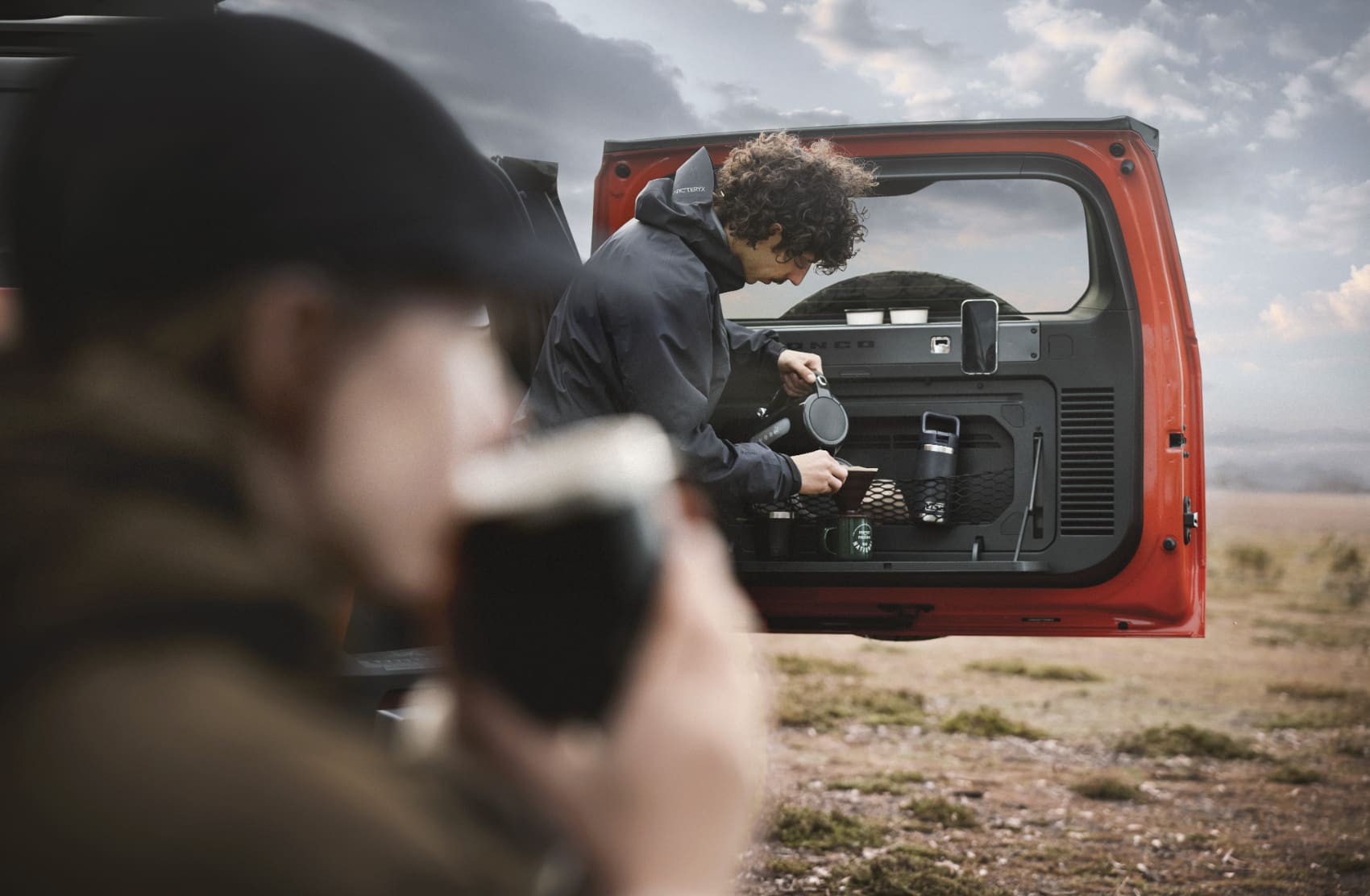 A person prepares a drink at the back of an open red Ford Bronco, while another person in the foreground drinks from a cup outdoors.