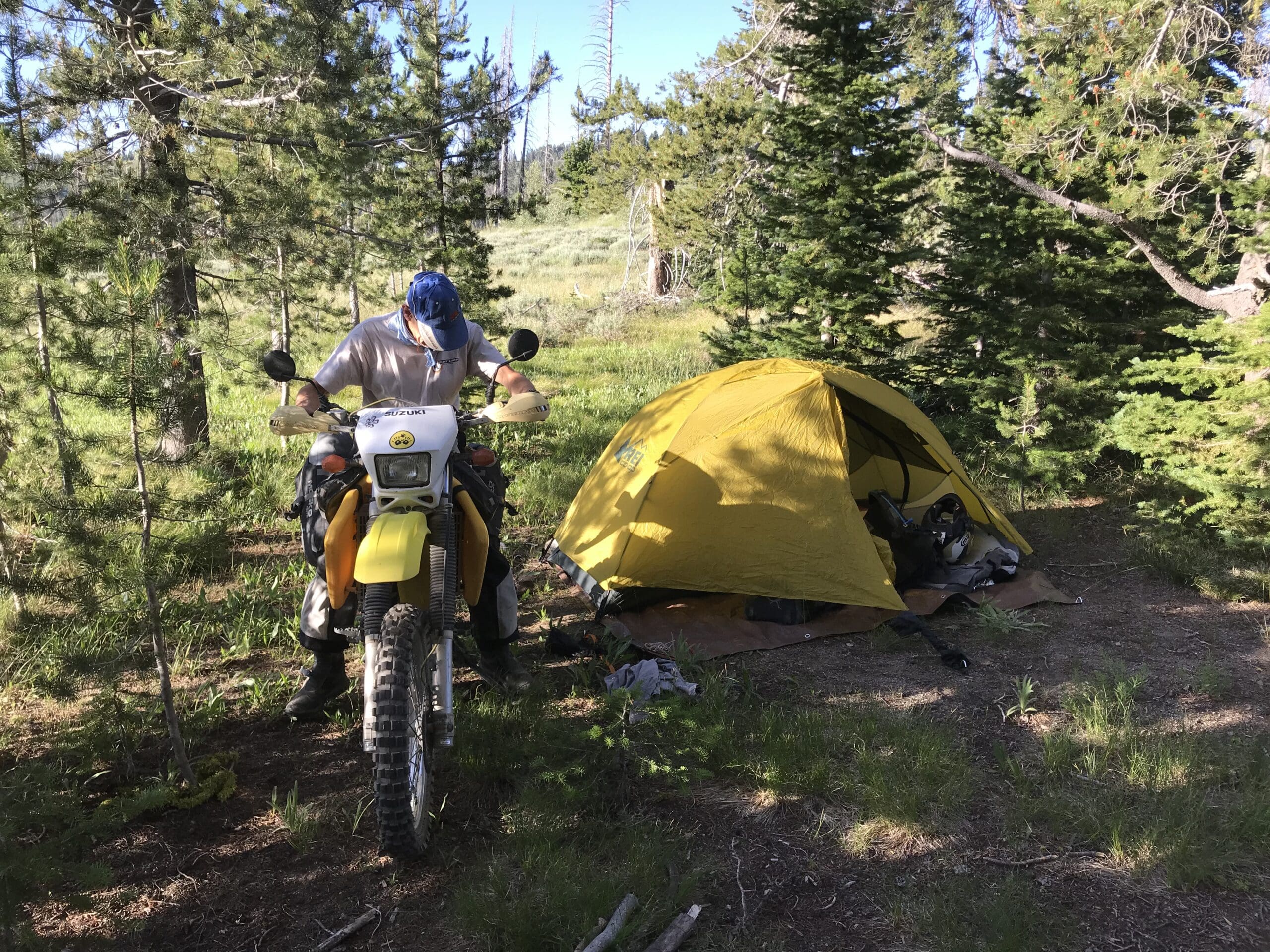A person wearing a helmet sits on a yellow dirt bike near a yellow tent in a forested campsite, surrounded by trees and sunlight filtering through the branches—a quiet moment that feels like a true Celebration of Life, reminiscent of Arden Kysely’s adventurous spirit.
