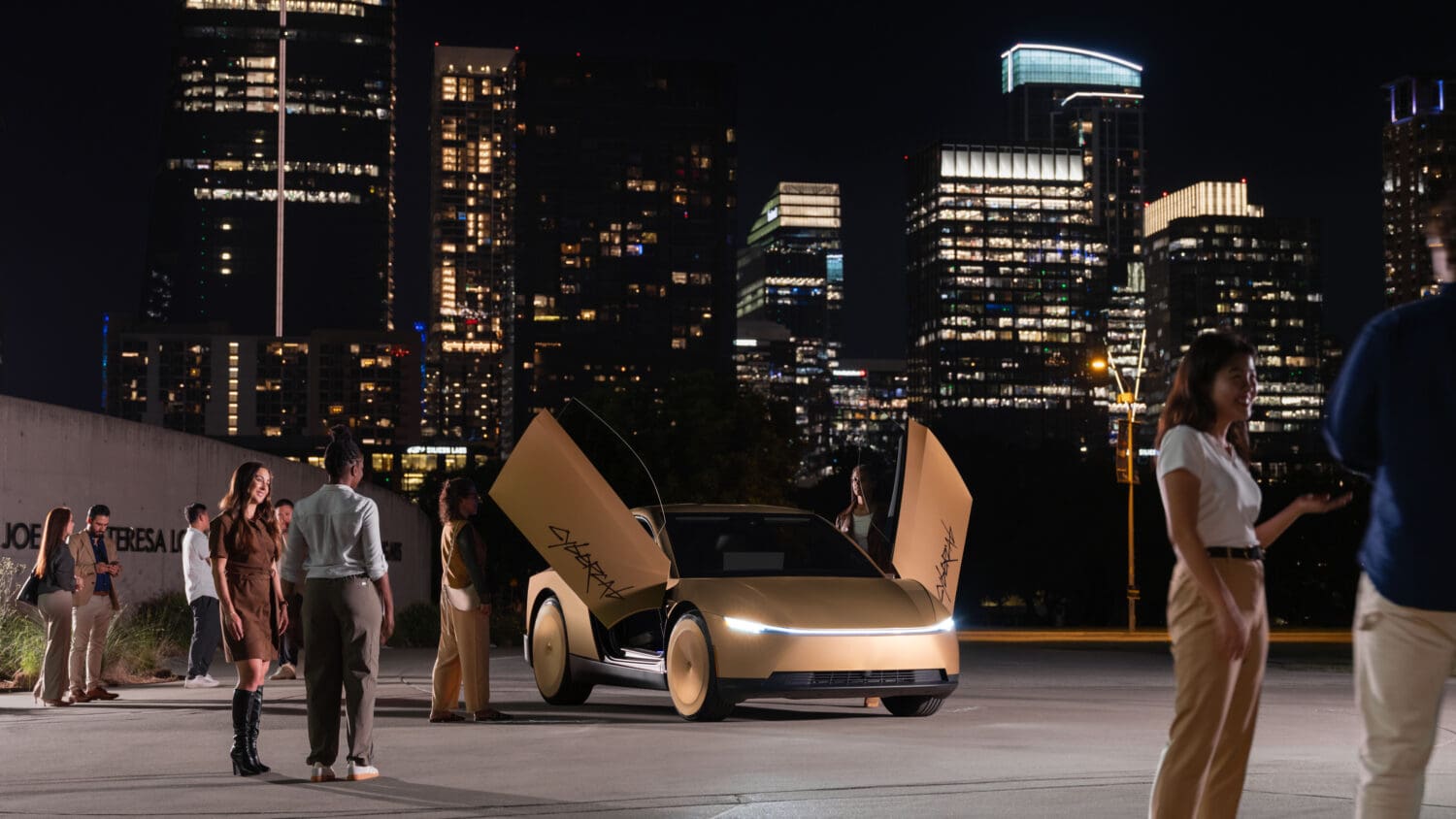 A gold concept car with open doors is surrounded by people at night, with tall city buildings lit up in the background, showcasing the future of autonomous overlanding.