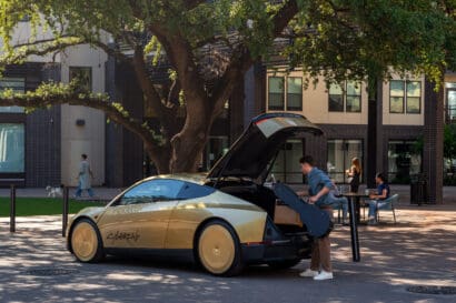 A person loads a guitar case into the trunk of a gold and black car with gullwing doors—an impressive example of autonomous overlanding—parked on a brick street in front of a modern apartment building and large tree. Other people are in the background.