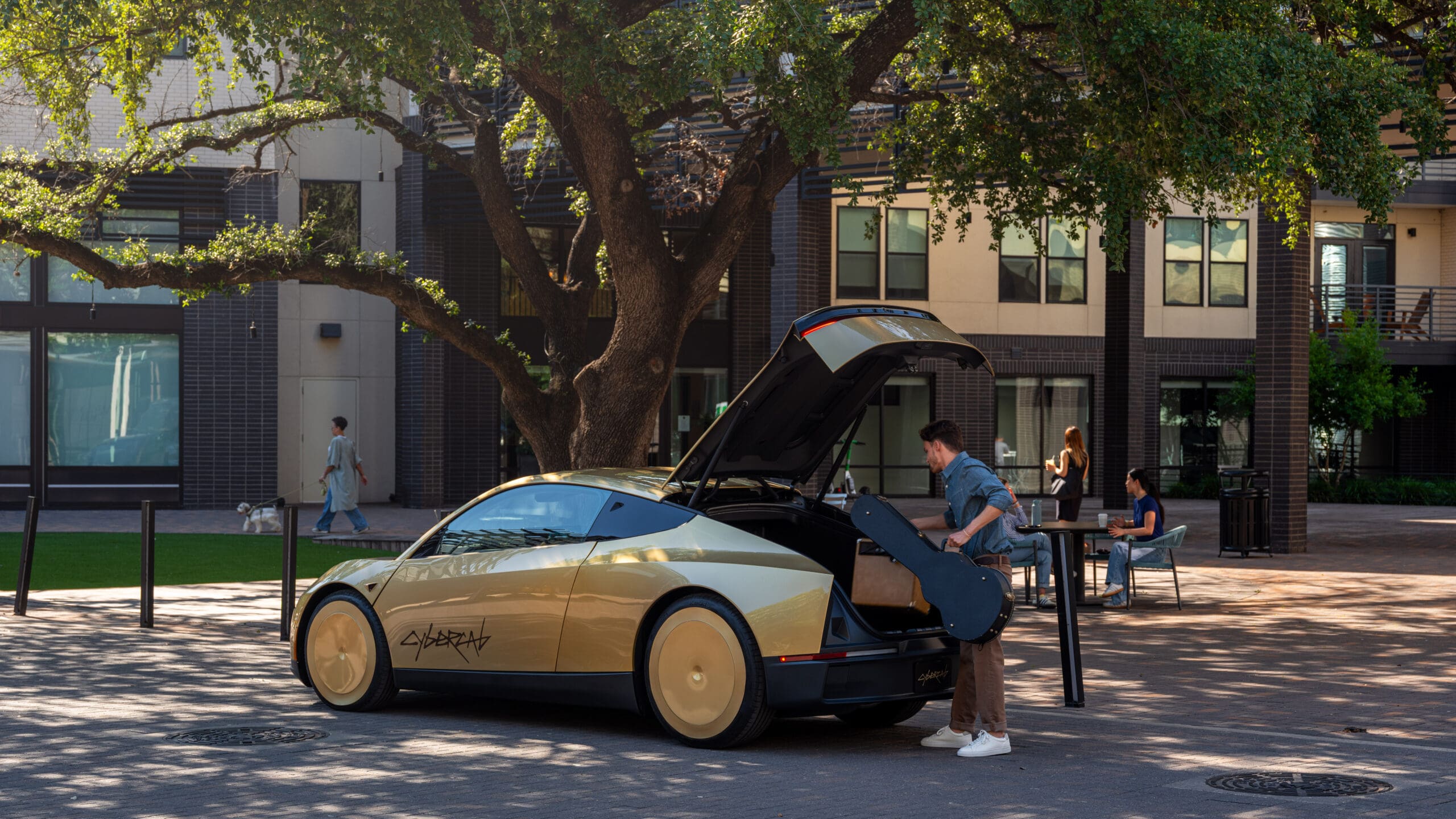 A person loads a guitar case into the trunk of a gold and black car with gullwing doors—an impressive example of autonomous overlanding—parked on a brick street in front of a modern apartment building and large tree. Other people are in the background.