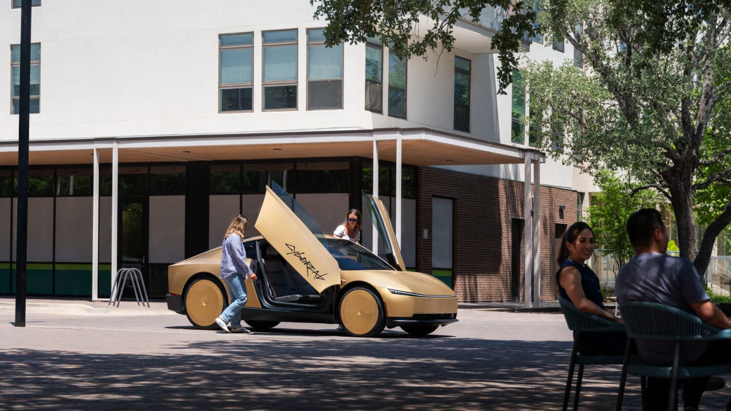 A gold car with upward-opening doors—perfect for autonomous overlanding adventures—is parked by a building. Two people stand near the car, while two others sit at a table nearby under trees.