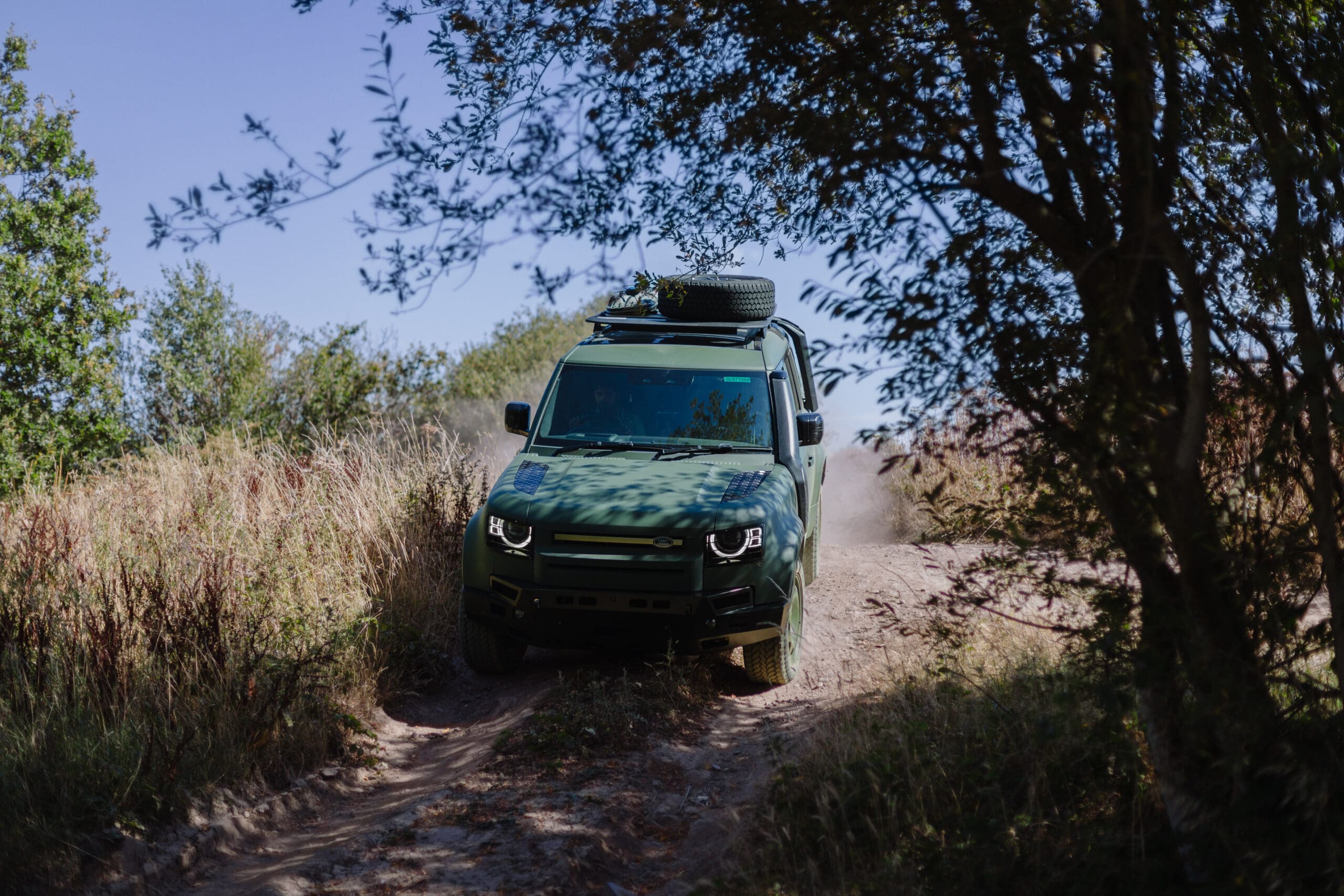 A green Defender Wolf Series II off-road vehicle drives down a dusty dirt path, surrounded by tall grass and trees under a clear blue sky.