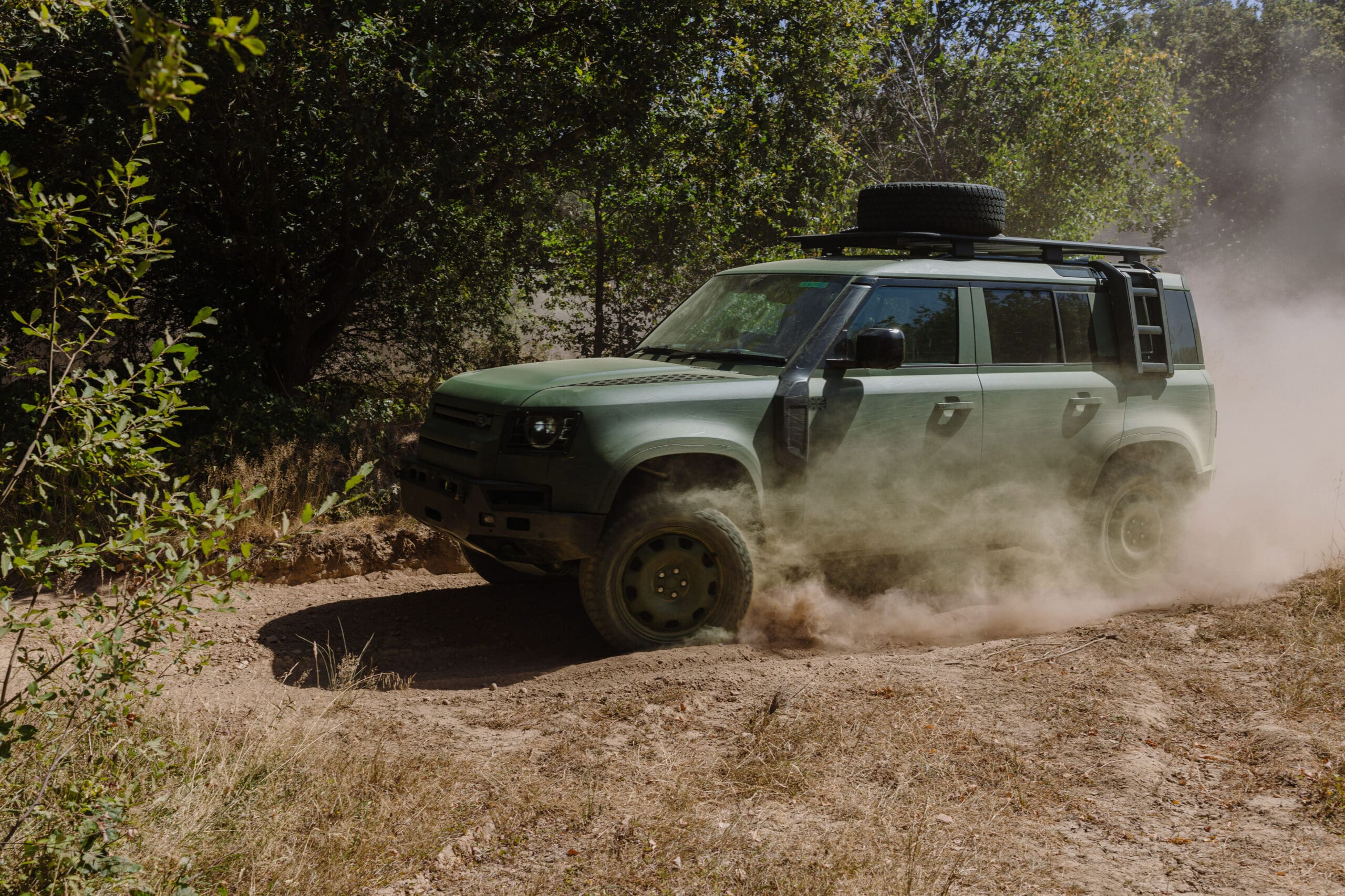 A green Defender Wolf Series II SUV drives on a dusty dirt road surrounded by trees, kicking up dust as it goes around a curve.
