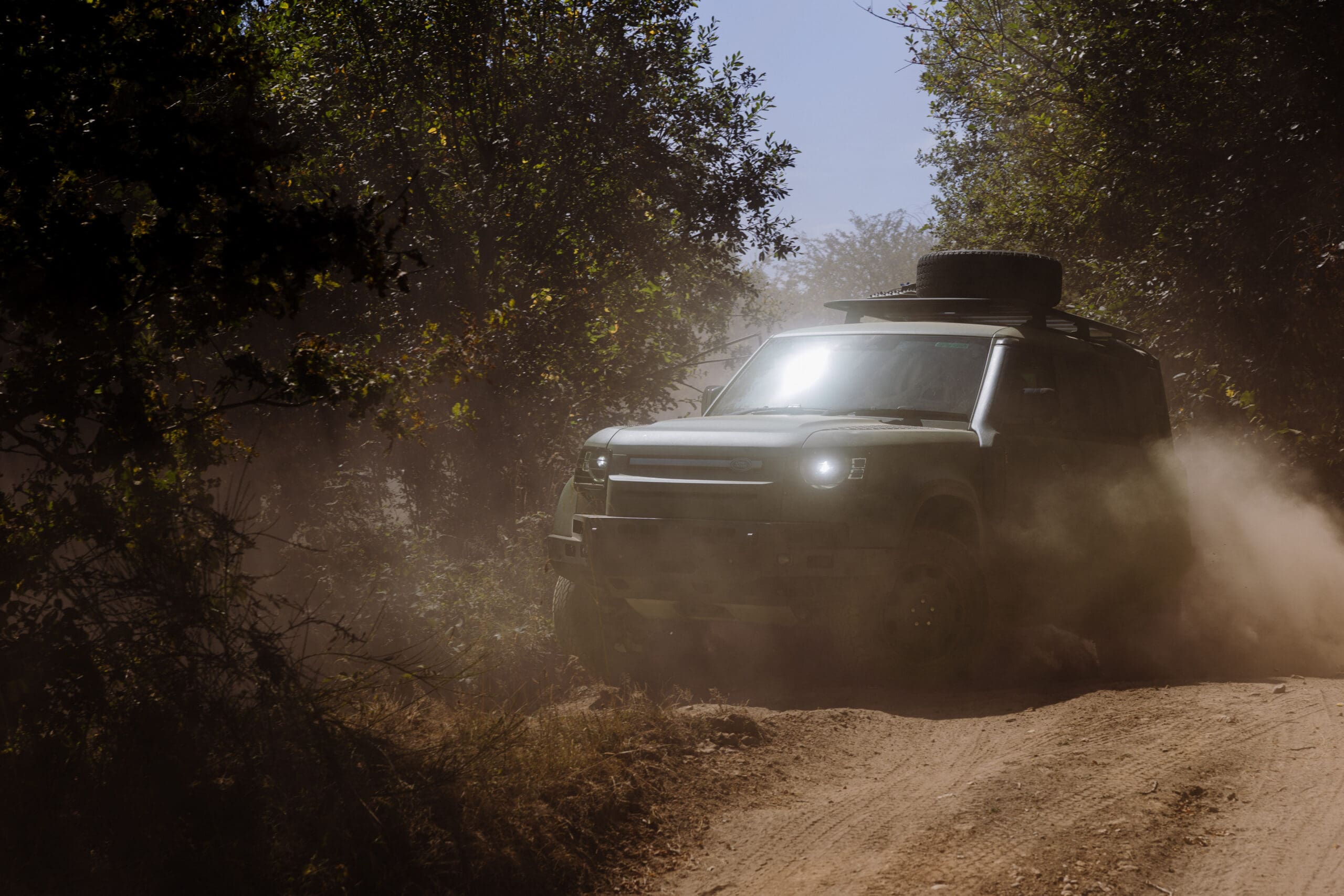 A rugged Defender Wolf Series II SUV kicks up dust as it drives through a narrow, dirt forest trail, surrounded by green trees and sunlight filtering through branches.