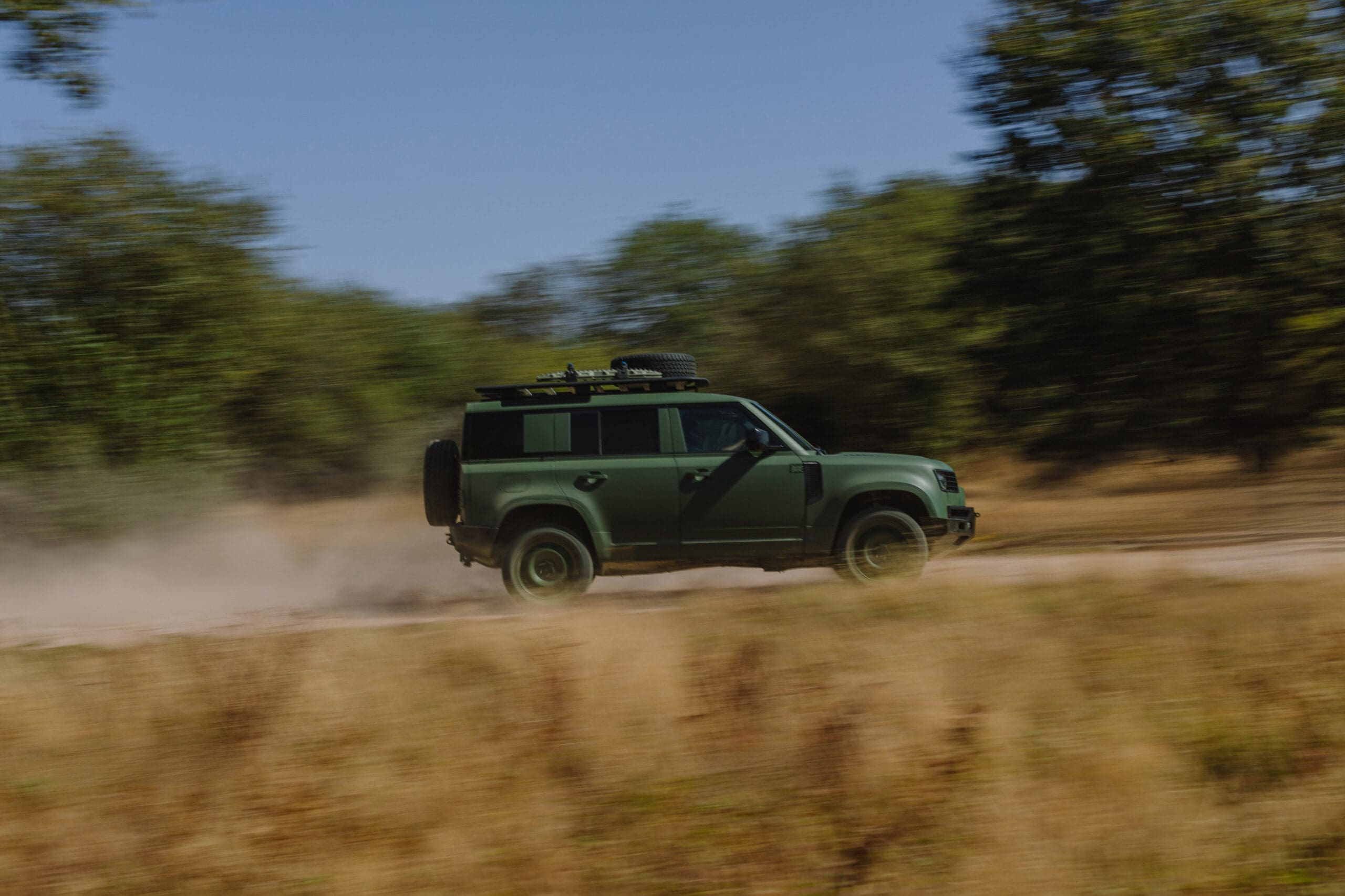 A green Defender Wolf Series II SUV speeds down a dusty dirt road, surrounded by dry grass and trees, with its roof rack loaded with gear.