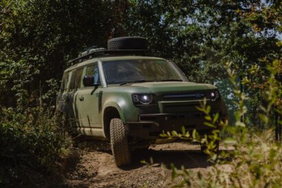 A green Defender Wolf Series II off-road vehicle drives on a dirt path surrounded by dense trees and bushes on a sunny day.