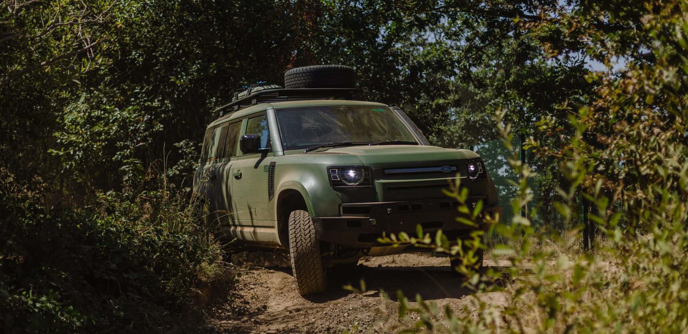 A green Defender Wolf Series II off-road vehicle drives on a dirt path surrounded by dense trees and bushes on a sunny day.