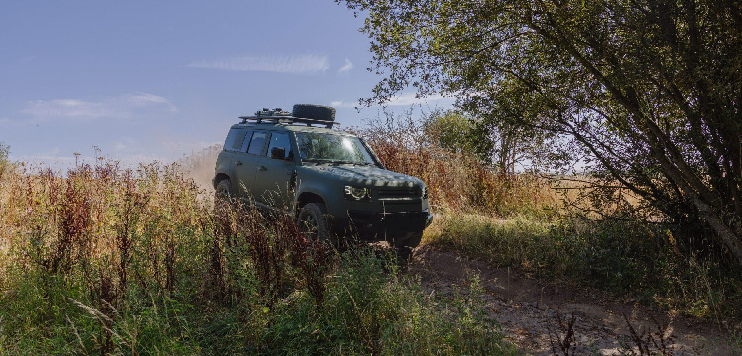 A Defender Wolf Series II green SUV drives off-road through tall grass near a tree under a clear blue sky, kicking up dust behind it.