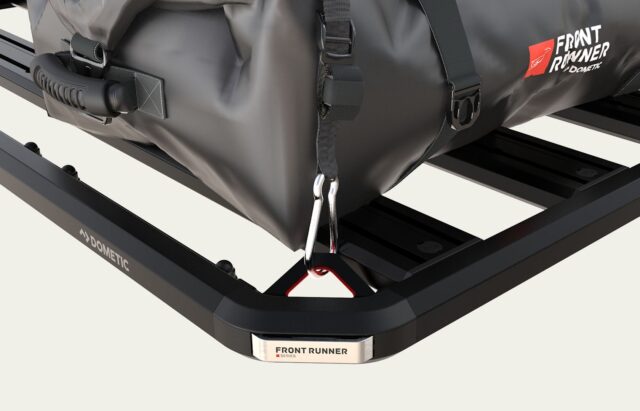 A close-up of a black roof rack with a gray duffel bag secured by a carabiner. The rack, featured in Overland News, displays Front Runner and Dometic labels visible on metal plates.