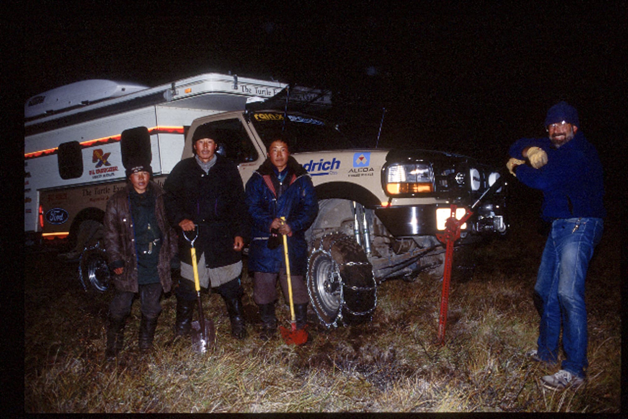 Four people stand in front of an off-road vehicle at night, holding shovels. Another person on the right is using a hi-lift tool to dig in the grassy terrain. The scene is lit by the vehicle’s headlights.