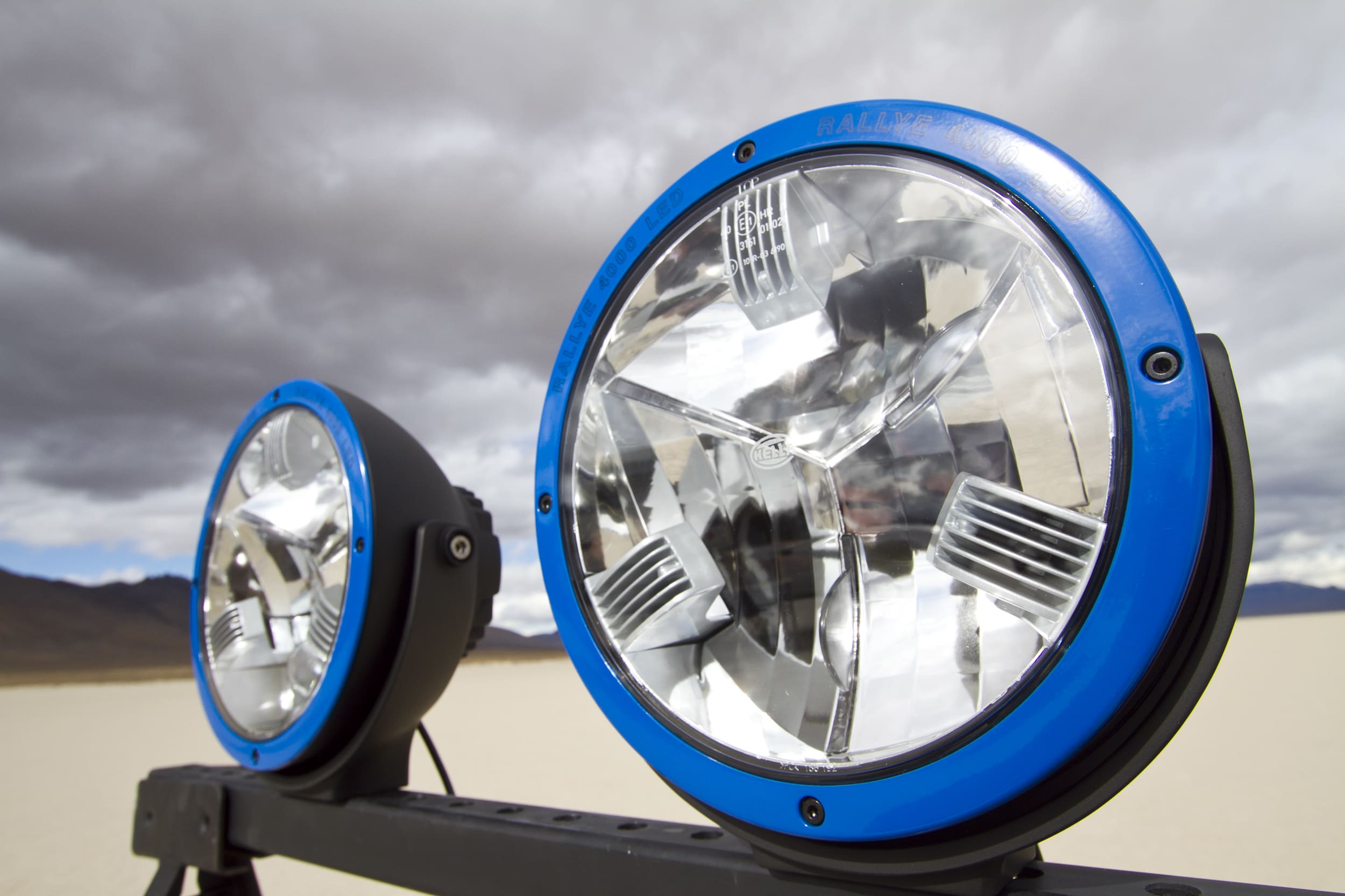 Two large round driving lights with blue rims are mounted on a metal bar outdoors, with a desert landscape and cloudy sky in the background.