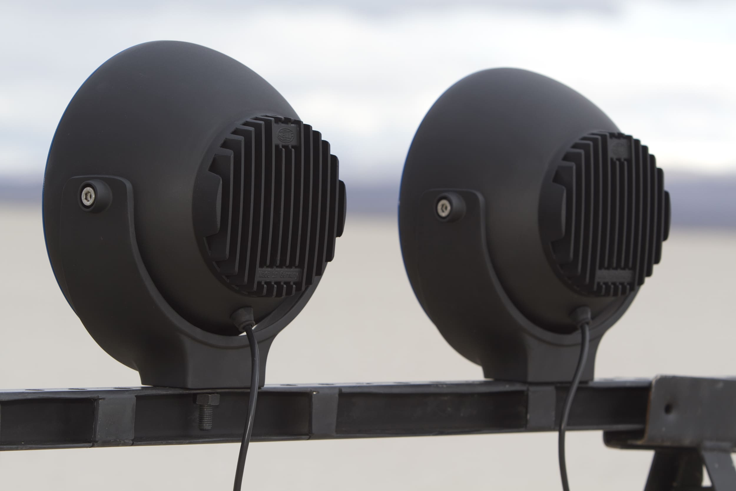 Two black, dome-shaped driving lights with heat sinks and visible power cords are mounted on a metal bar against a blurred outdoor background.