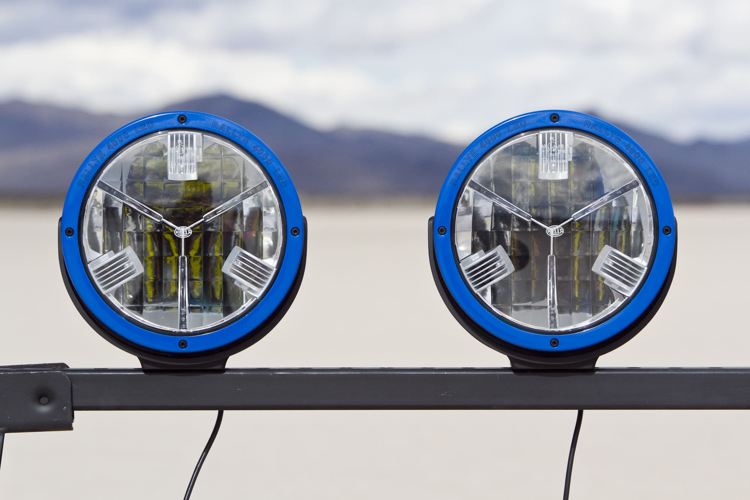 Two round, blue-rimmed driving lights are mounted on a metal bar outdoors, with mountains and a cloudy sky blurred in the background.