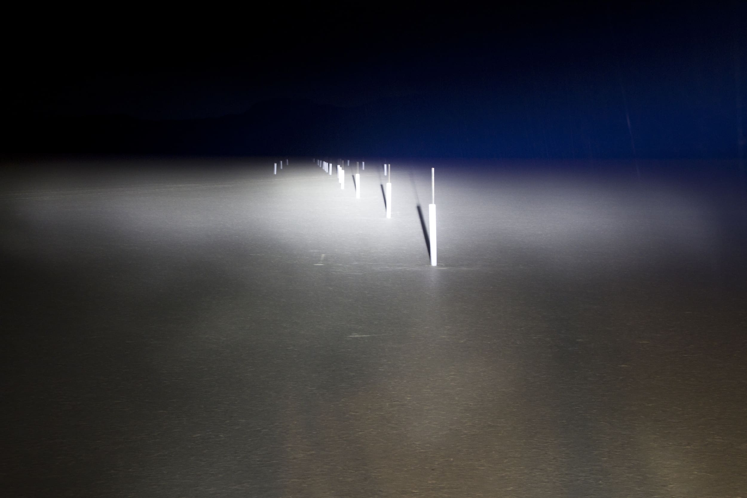 A dark road is dimly lit by driving lights, revealing a row of white reflective road markers extending into the distance. The scene is shrouded in darkness and fog.