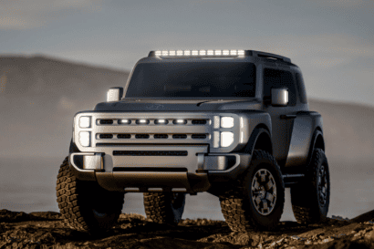 A rugged, futuristic Hyundai SUV with large tires and LED lights sits on rocky terrain near water, with hills in the background under a cloudy sky.