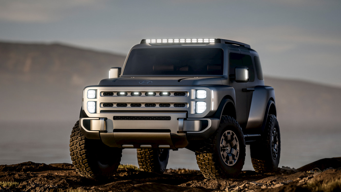 A rugged, futuristic Hyundai SUV with large tires and LED lights sits on rocky terrain near water, with hills in the background under a cloudy sky.
