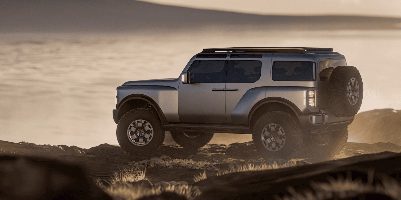 A rugged silver Hyundai SUV with a spare tire on the back drives off-road over rocky terrain at sunset, with a hazy landscape and water in the background.
