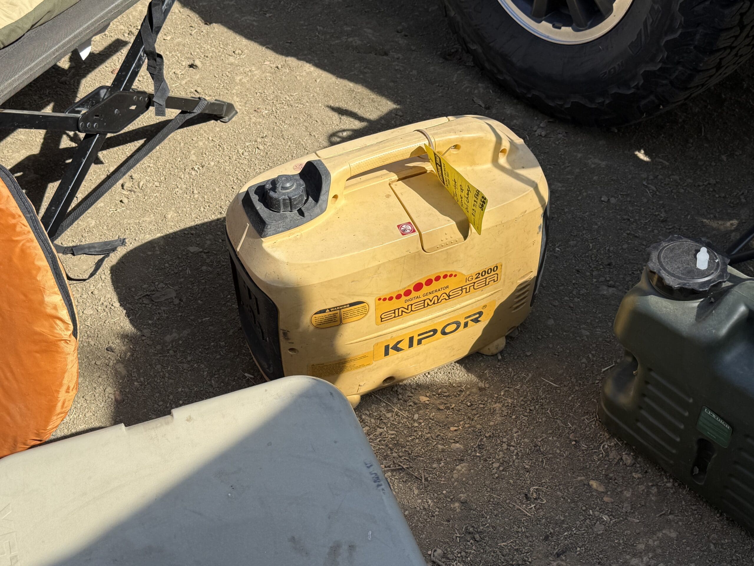 A yellow Kipor Enemaster generator sits on the ground outdoors next to camping gear, including a cot, a green container, and a 2011 Ford Ranger parked nearby in sunlight.