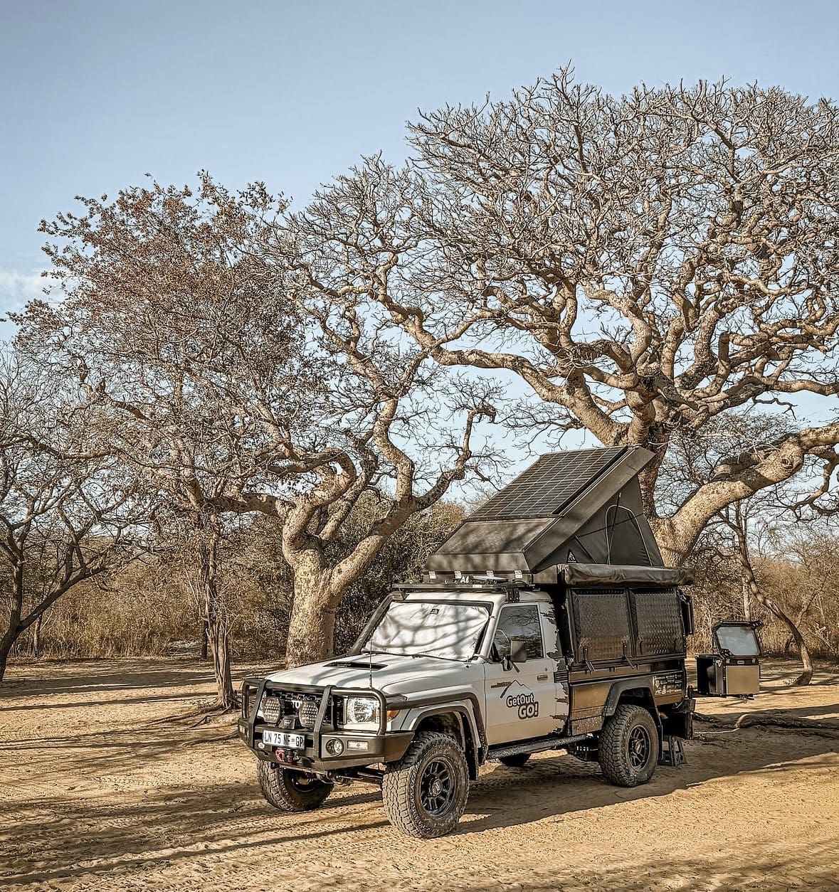 Get Out, Go! A rugged off-road camper truck with a pop-up roof is parked under large leafless trees on a sunny day in a dry, open landscape, ready for you to Get Out go exploring wherever the adventure leads.