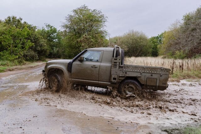 A muddy Defender Wolf Series II pickup truck drives through a large puddle on a rural dirt road, splashing water and mud, surrounded by grass and trees.
