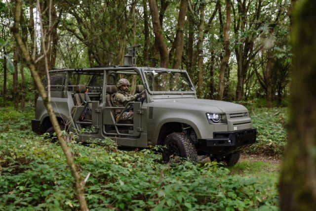 A Defender Wolf Series II military vehicle with an open frame drives through a dense, green forest. A person in camouflage gear is seated inside, surrounded by tall trees and thick underbrush.