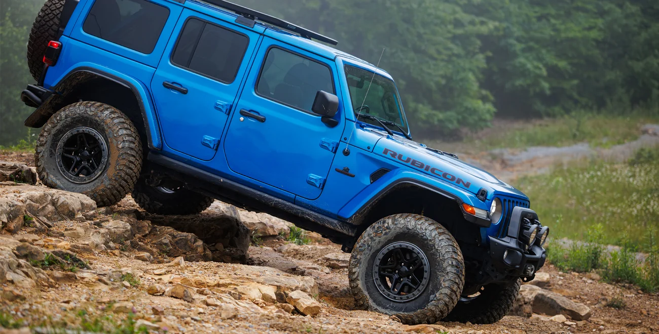 A blue Jeep Wrangler Rubicon off-roads on rocky terrain, angled upward with its front wheels elevated. Green trees and mist are visible in the background, setting the scene for this adventurous highlight featured in Overland News: News of the Week, September 2026.