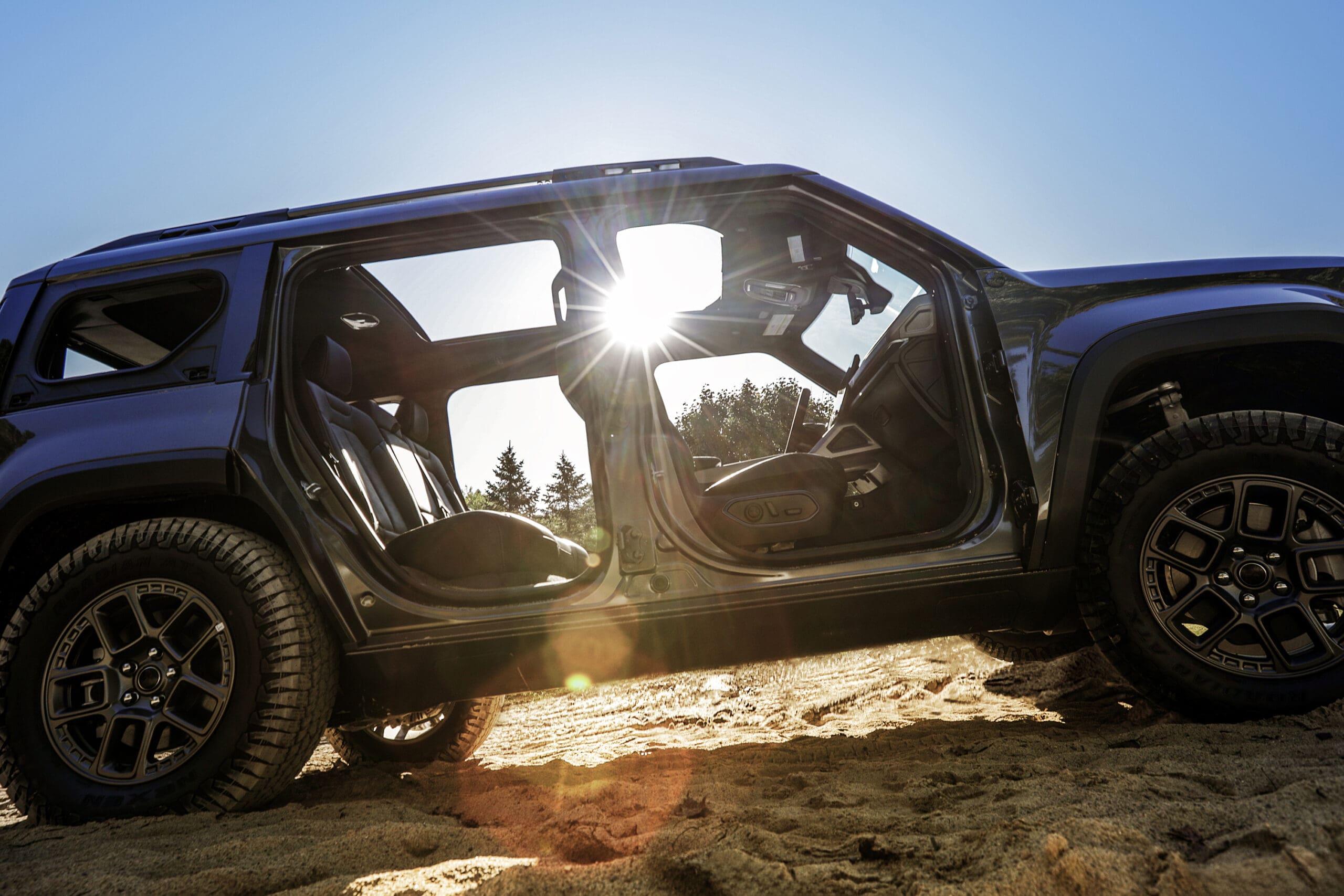 A black Jeep Recon SUV with no doors is parked on sand under a bright blue sky, with sunlight shining through the open frame and trees visible in the background.