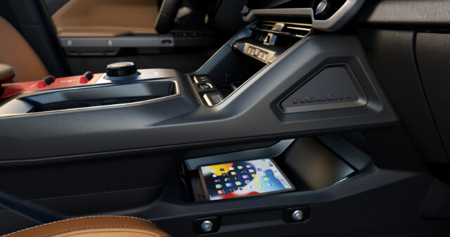 Close-up of a Jeep Recon’s front center console showing storage space with a tablet inside, various control buttons, and a brown leather seat partially visible.