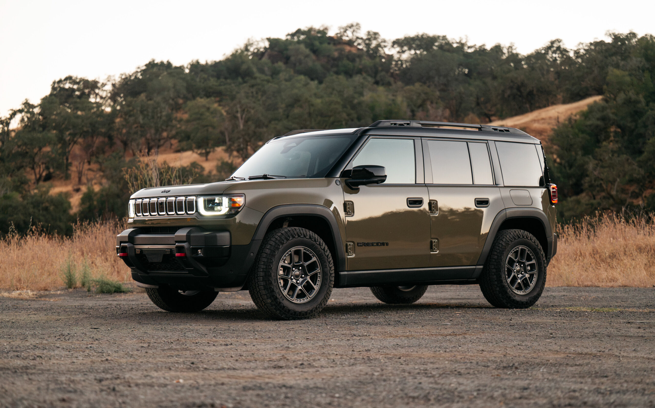 A metallic green Jeep Recon SUV with rugged tires is parked on a dirt surface with dry grass and trees in the background.