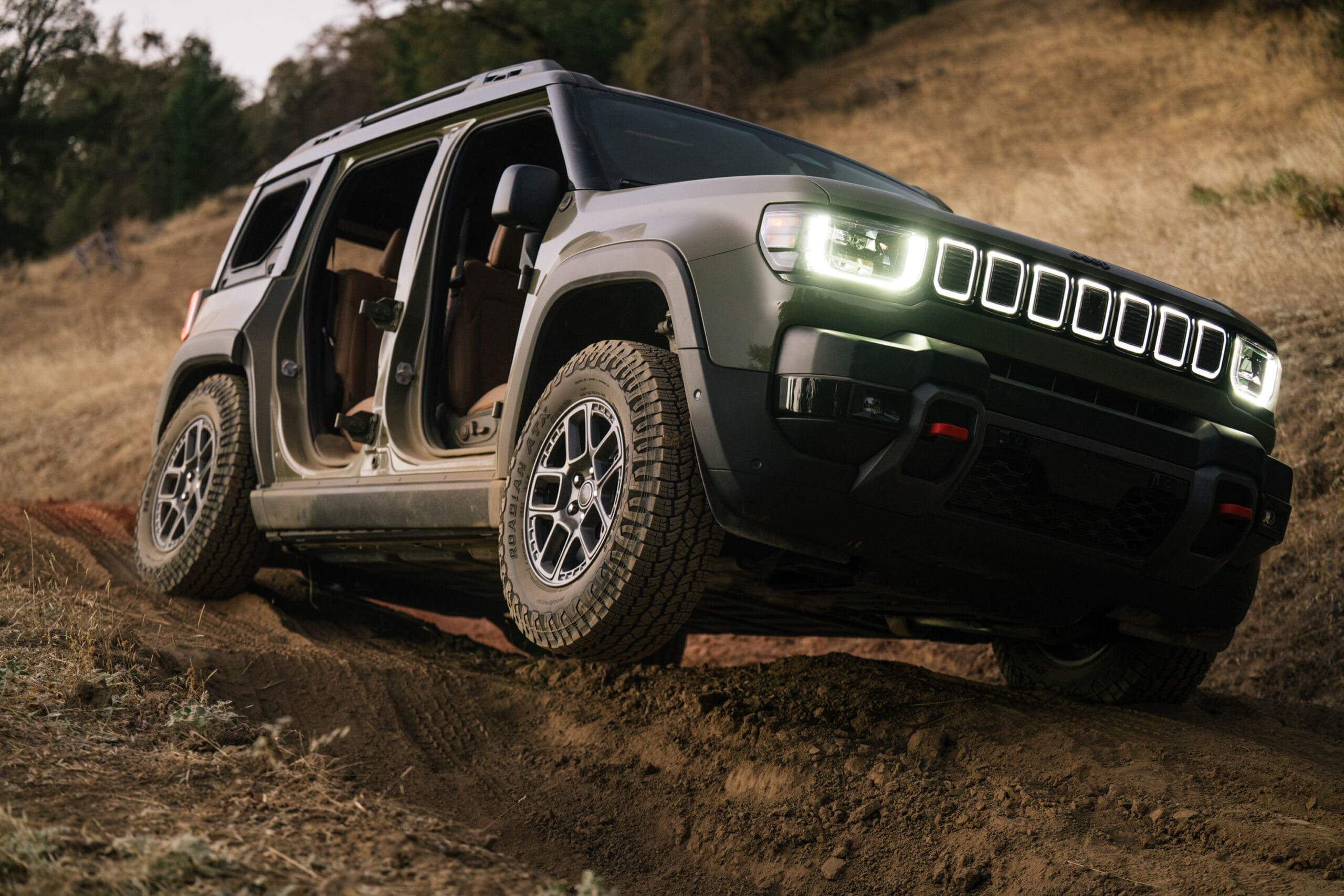 A rugged Jeep Recon SUV with its doors removed drives off-road on a dirt trail, headlights on, set against a dry, grassy hillside.