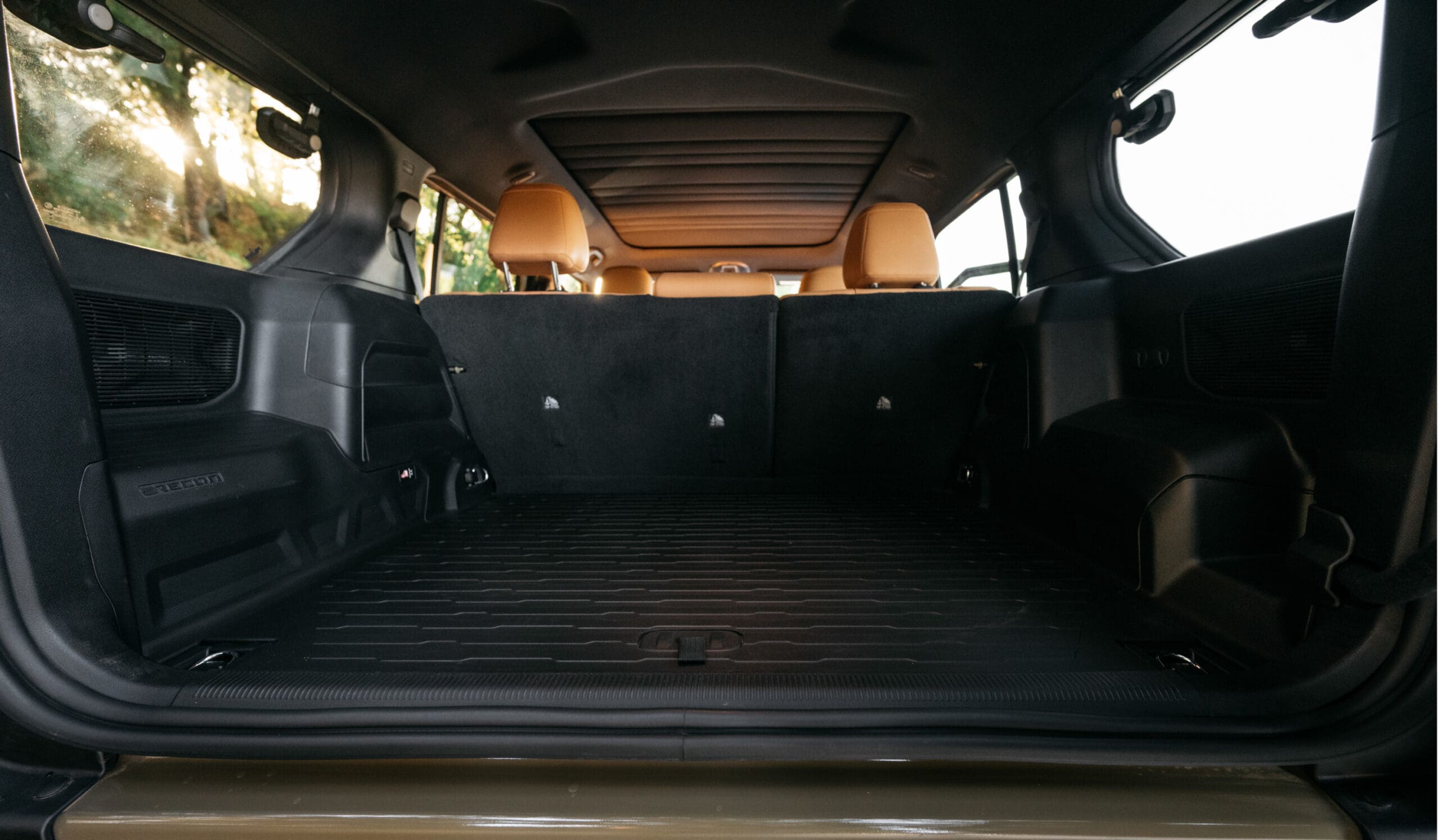 The image shows the spacious cargo area of a Jeep Recon SUV with the rear seats upright, featuring a black rubber mat and tan headrests in the back seats. Sunlight is visible outside the side window.