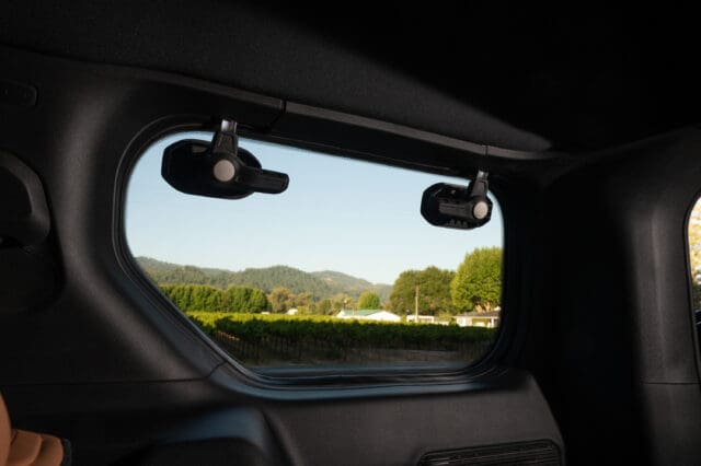 View through a Jeep Recon rear window showing a landscape with trees, hills, and clear sky. The window features two latches at the top.