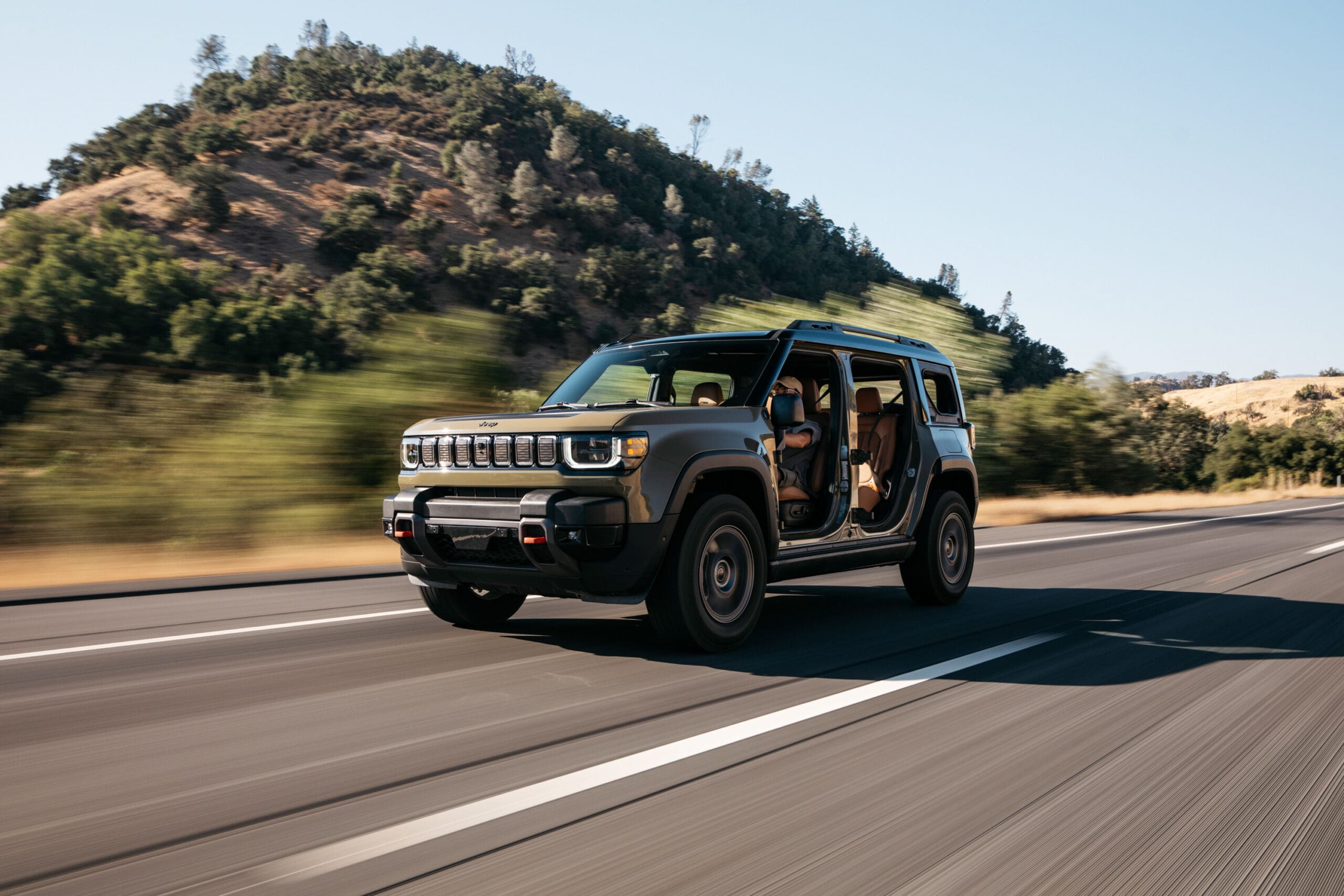 A gray Jeep Recon SUV with no doors or roof panels drives on a highway in a rural area, with a hillside and trees in the background under a clear sky.