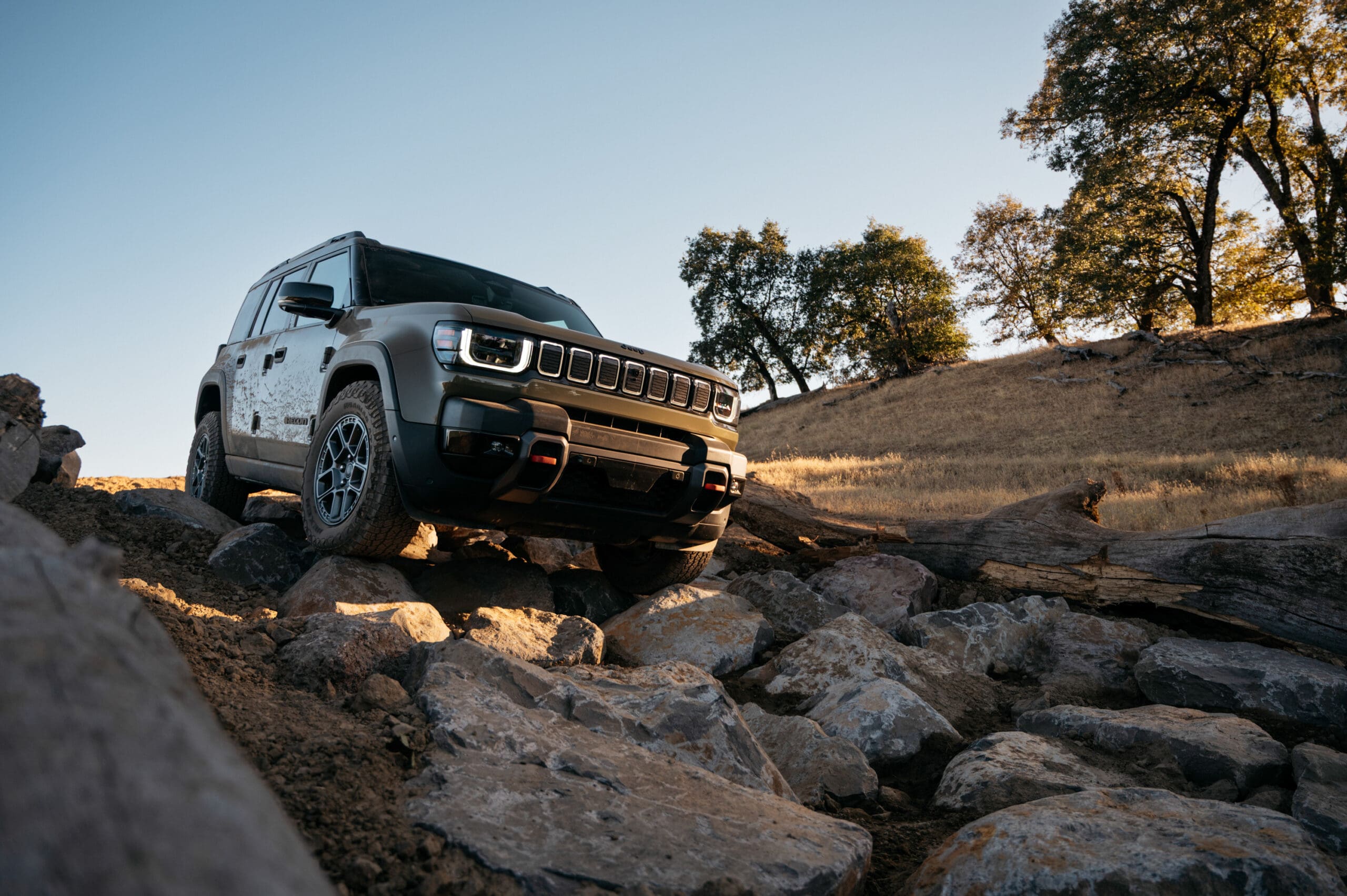 A rugged Jeep Recon SUV drives over large rocks and uneven terrain outdoors, surrounded by dry grass, fallen logs, and a few leafy trees under a clear blue sky.