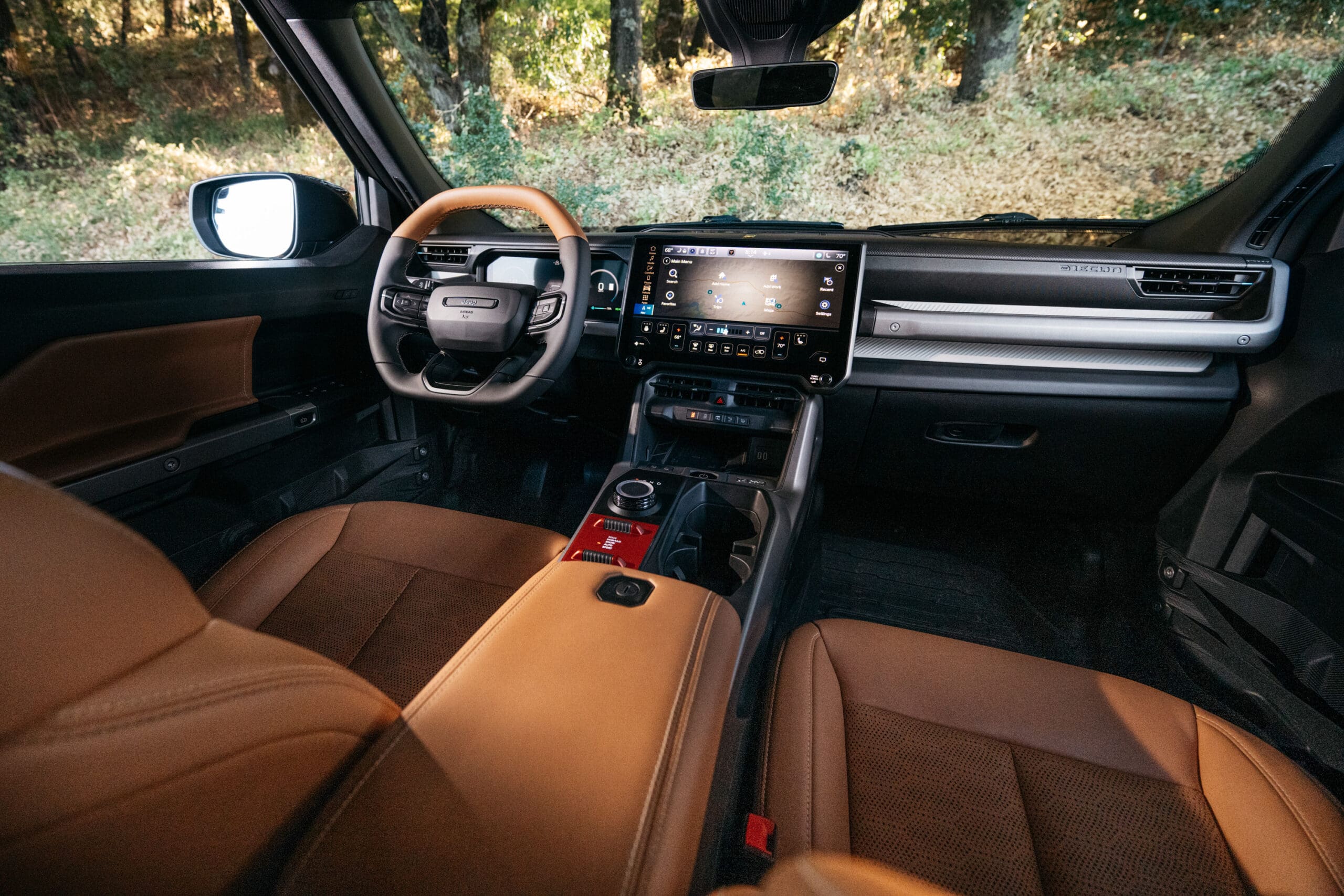 View of a modern Jeep Recon interior featuring brown leather seats, a digital dashboard, touchscreen display, and a rotary gear shifter, with trees visible through the windshield.