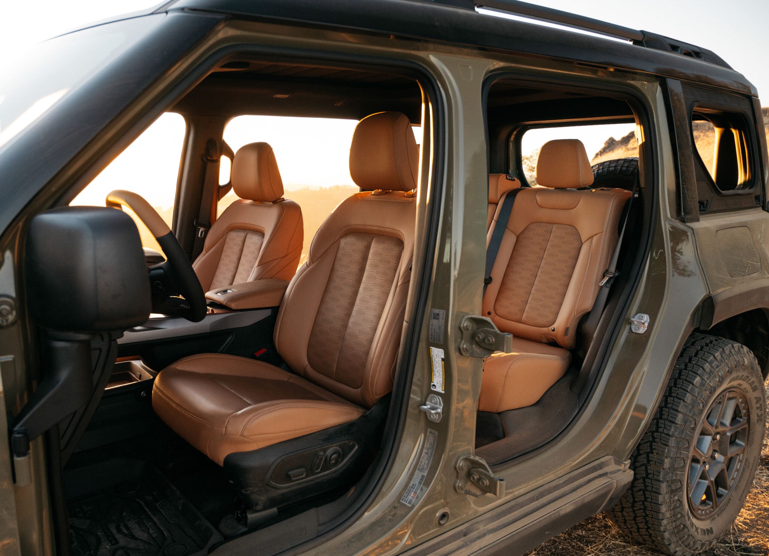A rugged Jeep Recon SUV with its doors removed, revealing tan leather interior seats. The vehicle is parked outdoors, with sunlight illuminating the cabin and dirt visible on the tires.