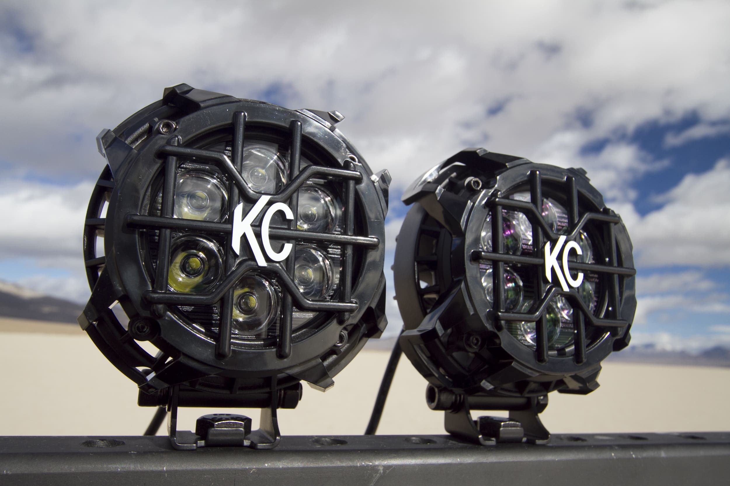 Two KC brand off-road driving lights with protective grills are mounted on a metal bar, set against a blurred desert landscape and cloudy sky in the background.