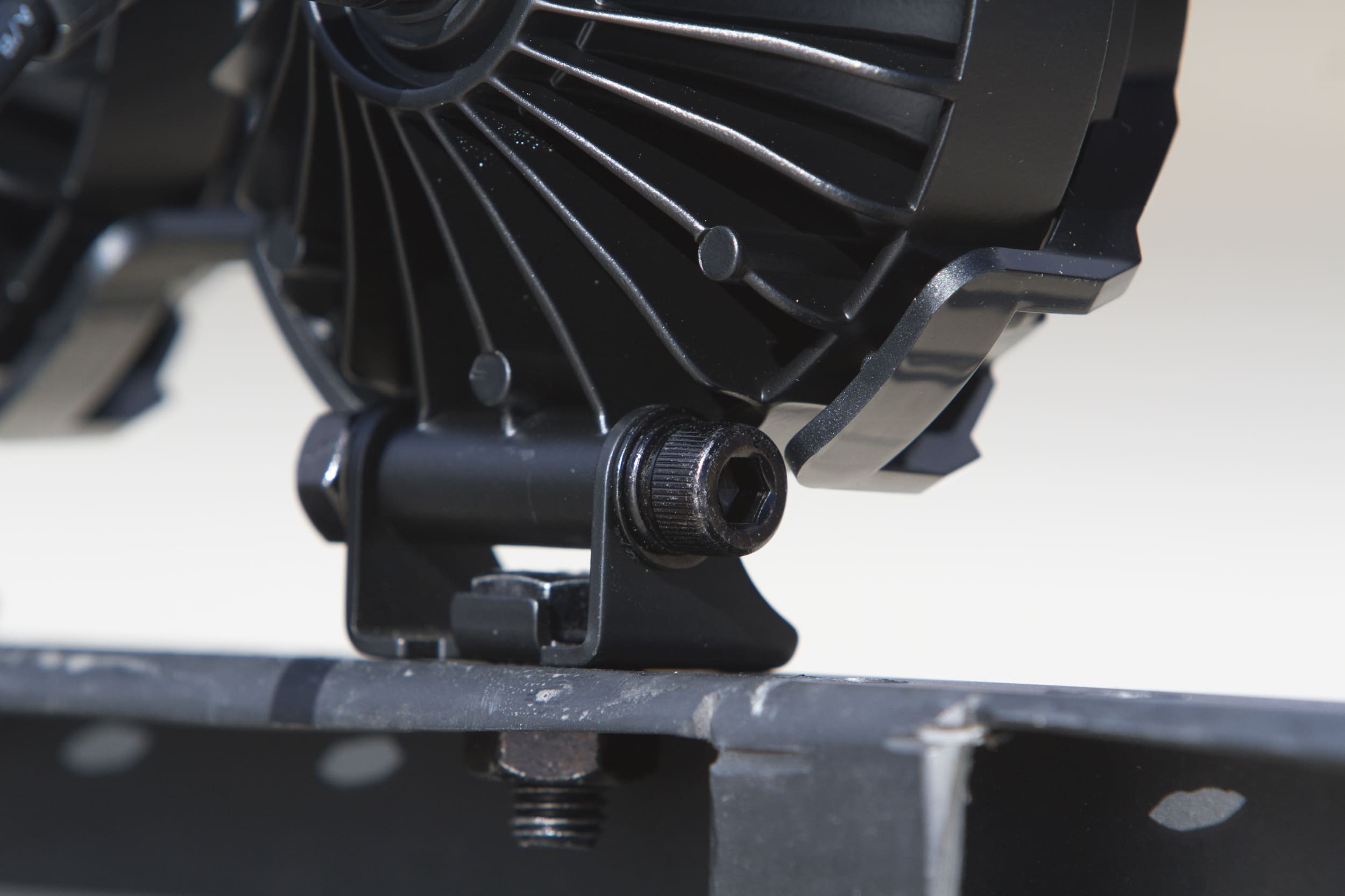 Close-up of a black metal mechanical component secured by a hex bolt and bracket, mounted on a perforated metal surface, with driving lights softly illuminating the blurred background.